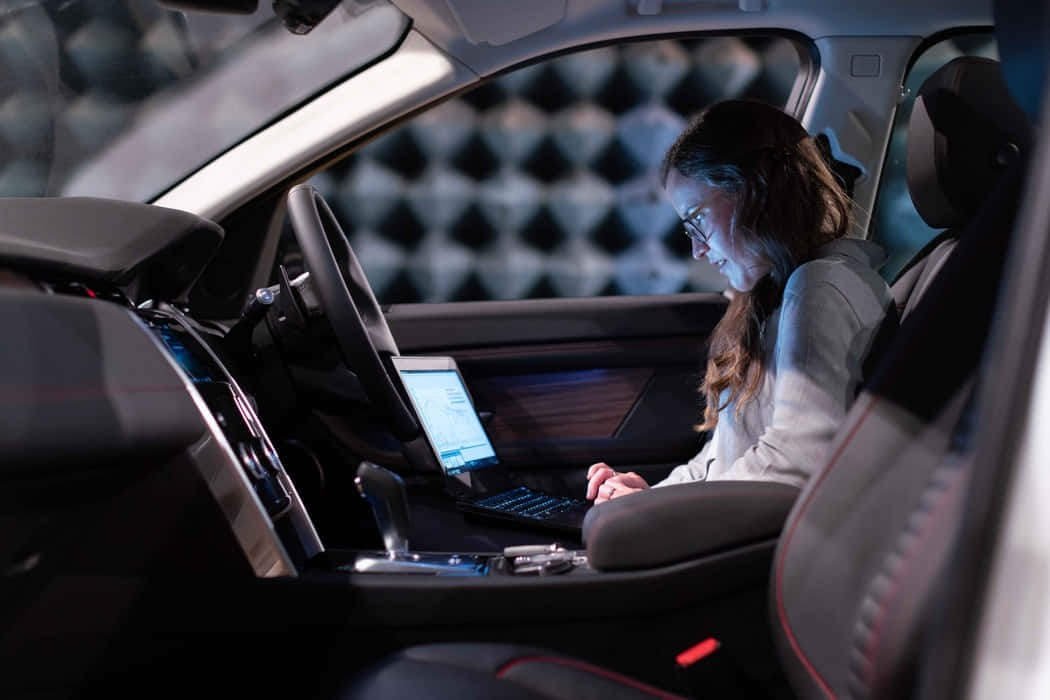 Car Technology 桌布 壁紙 — 一位戴眼鏡的女性在夜間於現代汽車內，在環境燈光下使用筆記型電腦工作。