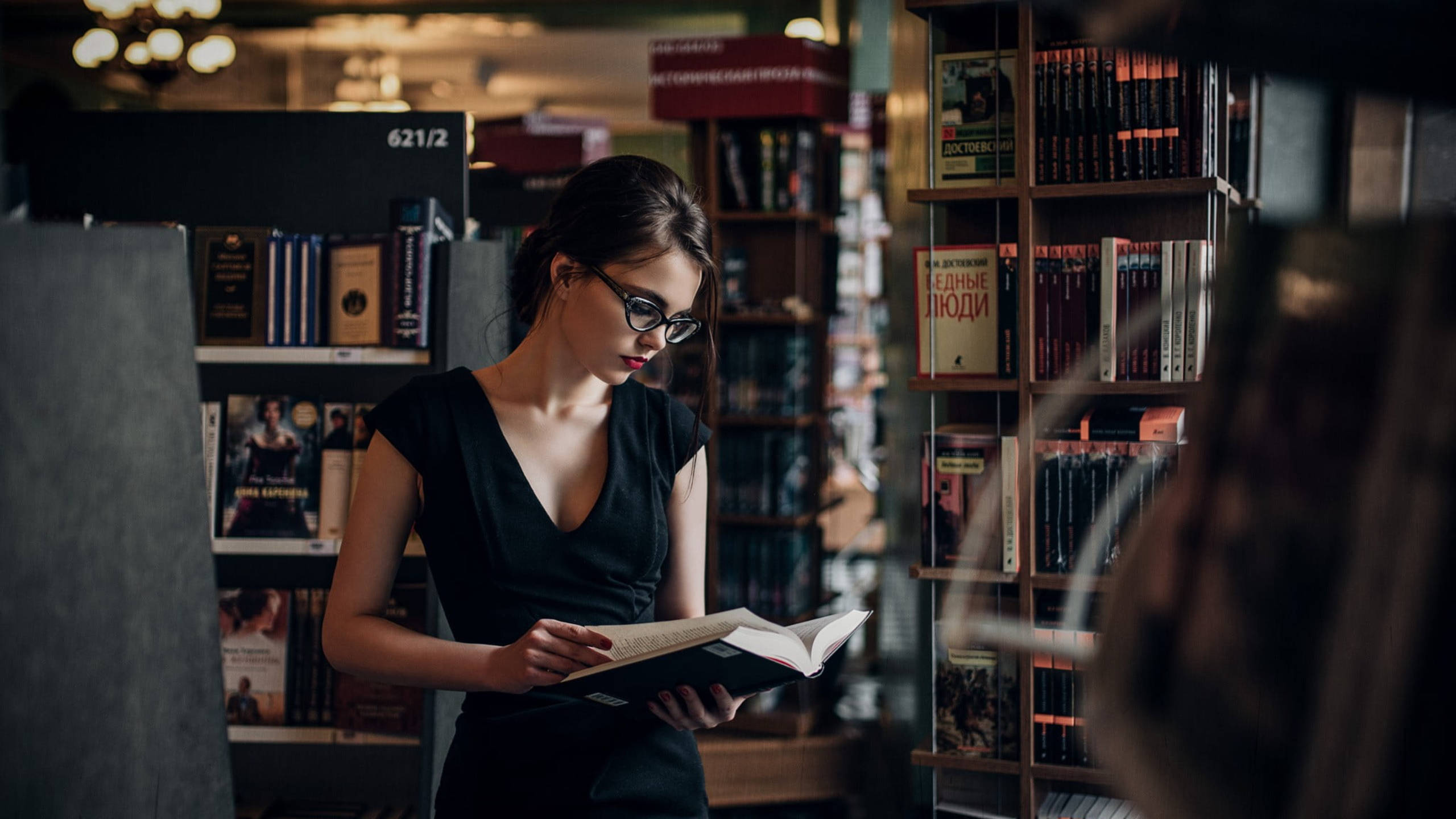 Reading 桌布 壁紙 — 一位戴眼鏡的女性在溫暖燈光的書店中閱讀，周圍是高高的書架。