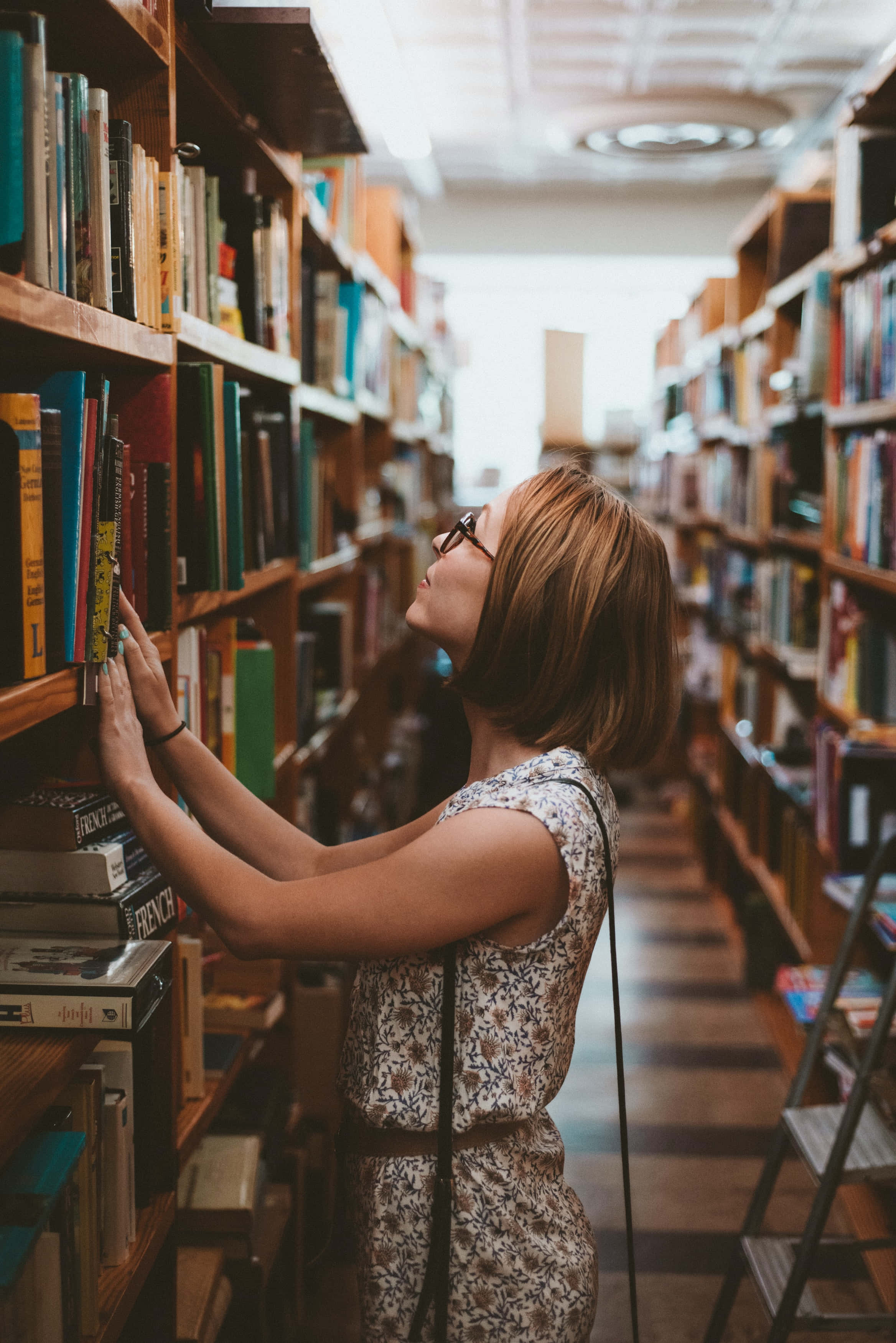Learning 桌布 壁紙 — 一位戴眼鏡的女性在燈光溫暖的圖書館走廊中，伸手去拿木書架上高處的書。