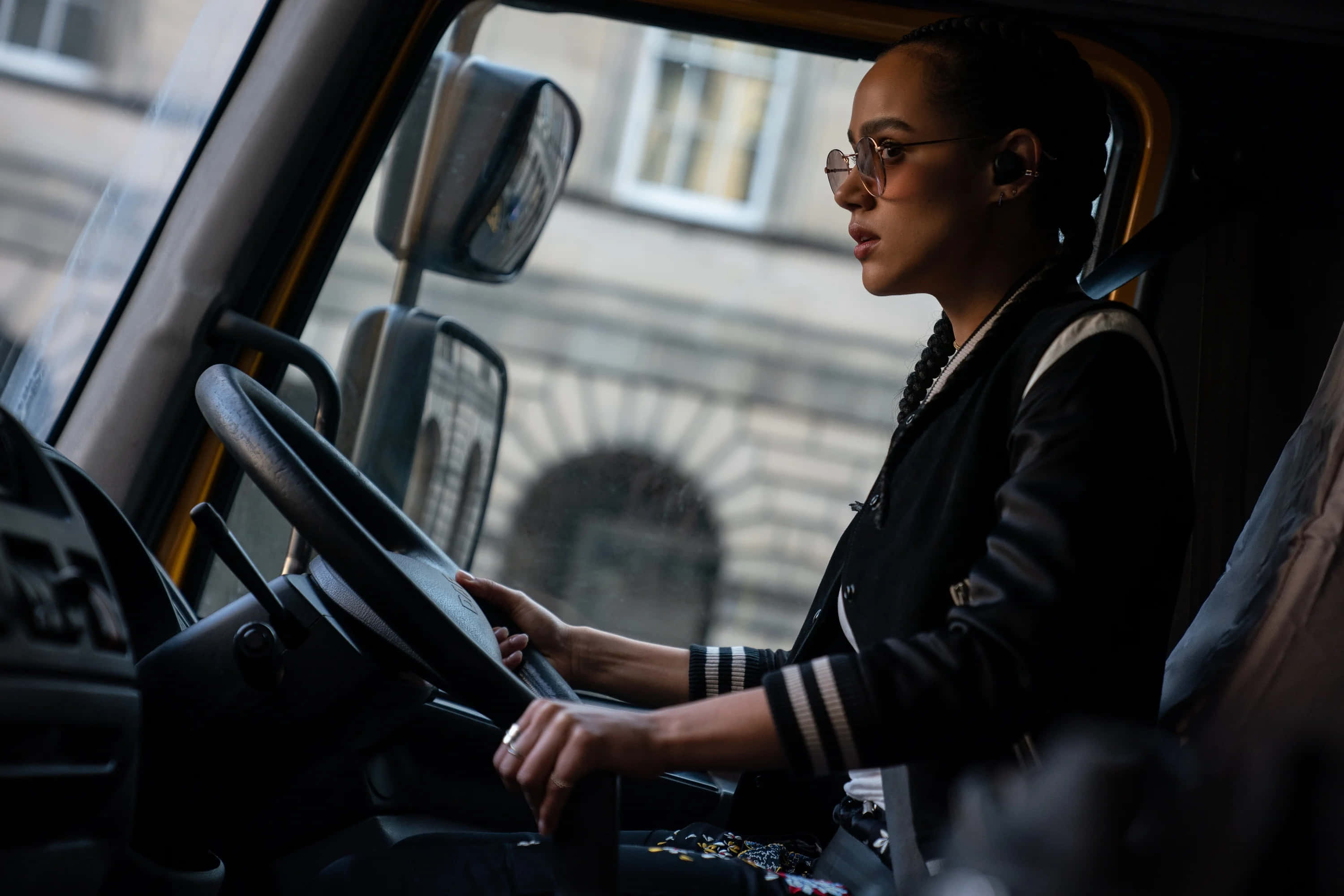 Fast And Furious 9 桌布 壁紙 — 一位戴眼鏡的專注女性在昏暗的駕駛室內握著貨車的方向盤，窗外可見城市建築。