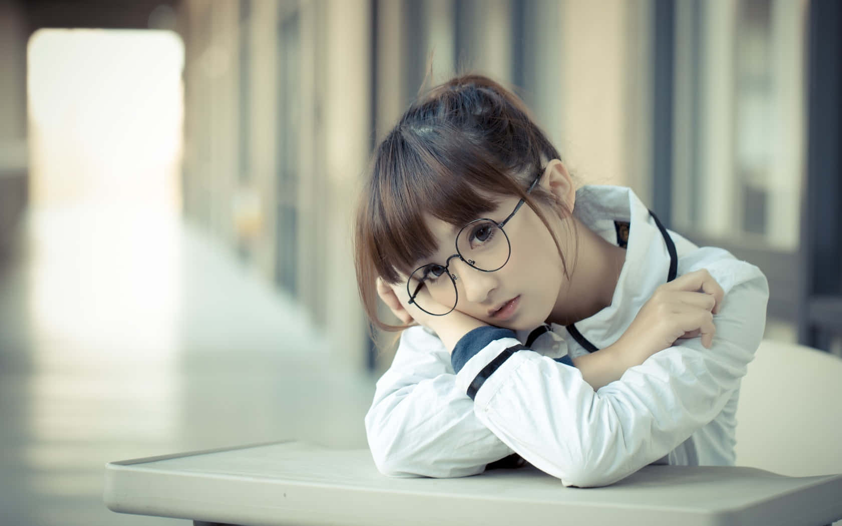 女孩 桌布 壁紙 — 一位戴眼鏡的年輕女子在光線柔和的走廊裡，將頭靠在桌上交叉的手臂上休息。
