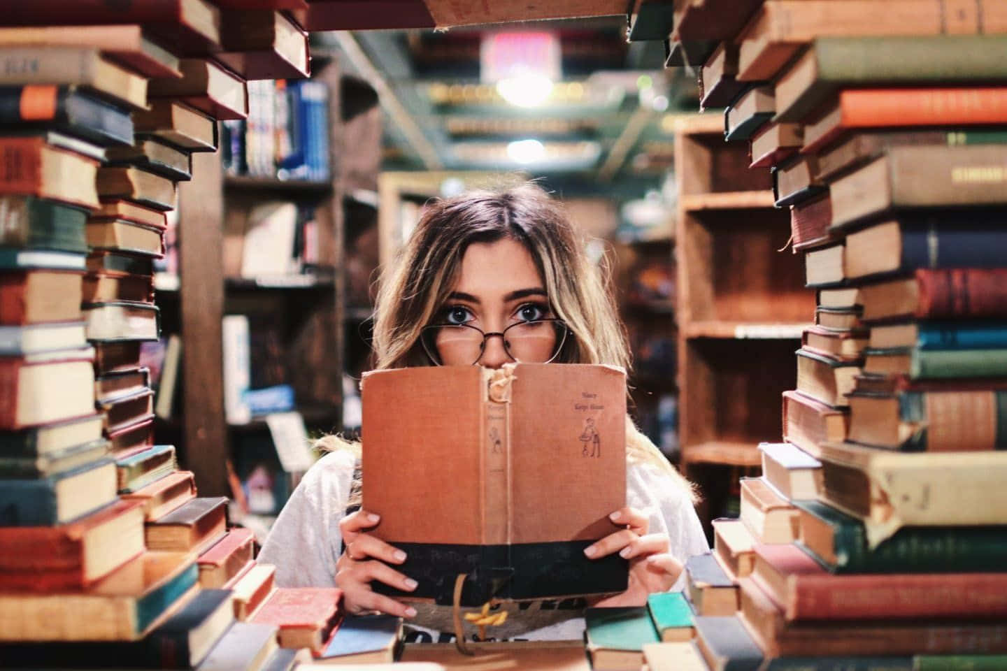 Library 桌布 壁紙 — 一位戴眼鏡的年輕女子從面前舉起的書本上方窺視，周圍是舒適圖書館中高聳的書堆。