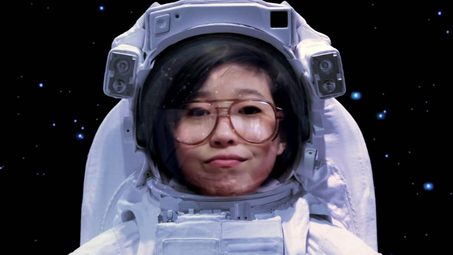 Awkwafina 桌布 壁紙 — 一位戴眼鏡的年輕女子身穿白色太空服，在繁星點點的黑色太空背景下平靜地凝視著鏡頭。