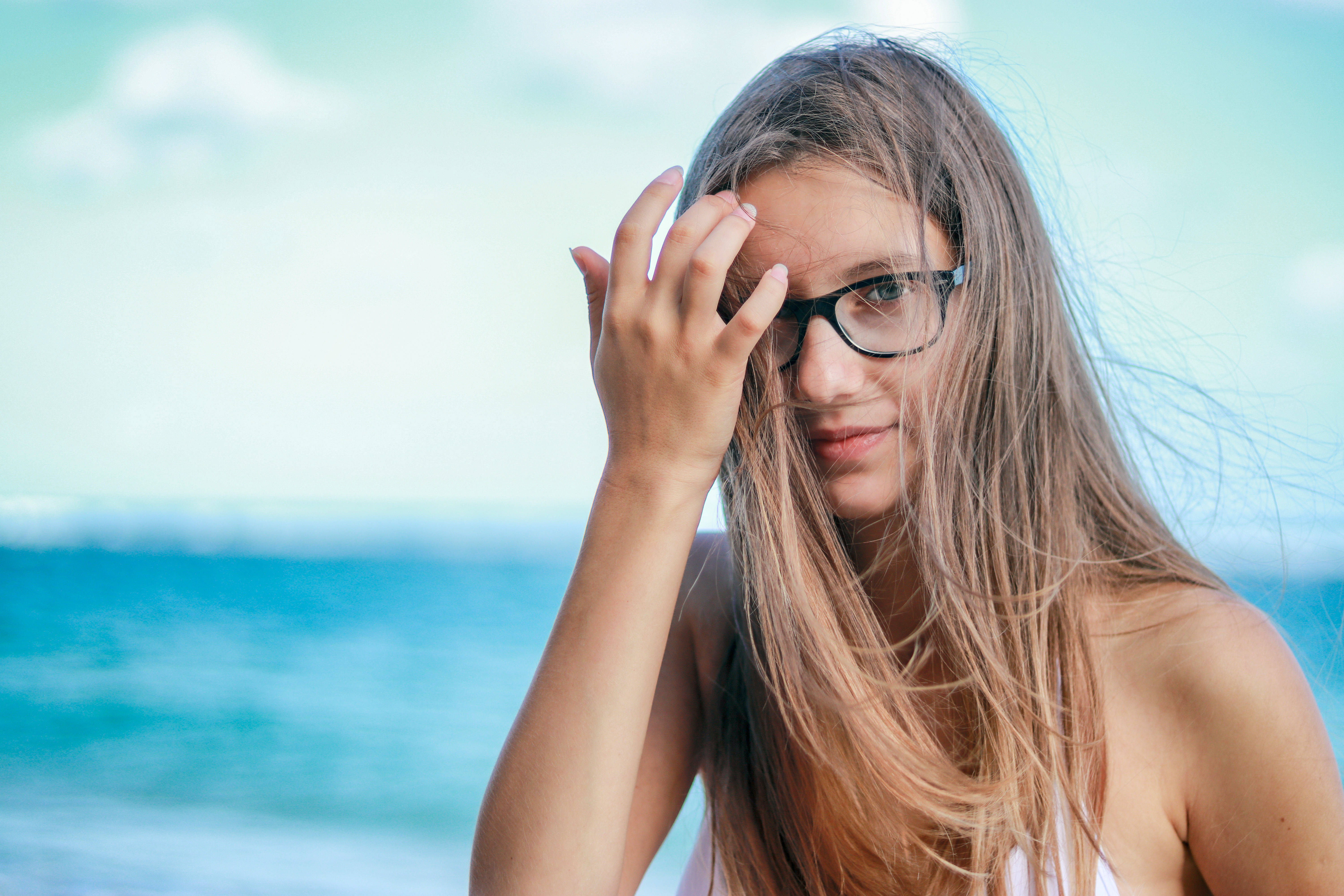 girls 桌布 壁紙 — 一位戴眼鏡的年輕女性，頭髮被風吹動，在明亮的天空下於海邊溫柔微笑。