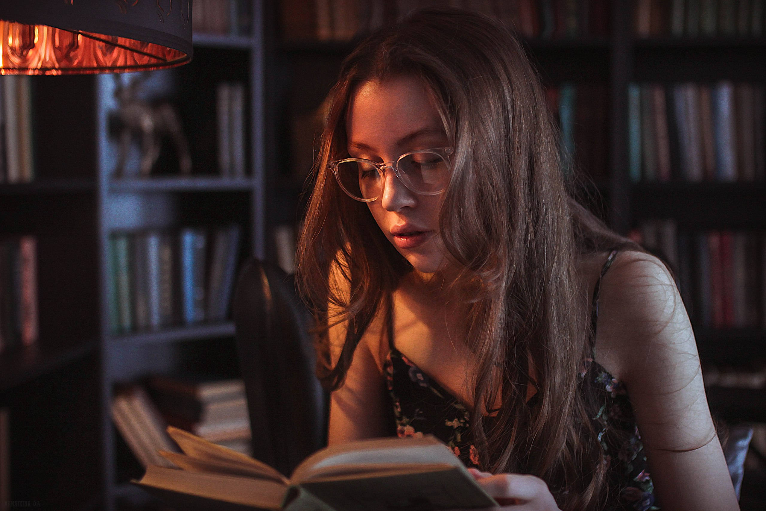 看書 桌布 壁紙 — 一位戴眼鏡的年輕女性在溫暖的燈光下，在一個充滿書架的舒適圖書館裡閱讀一本書。