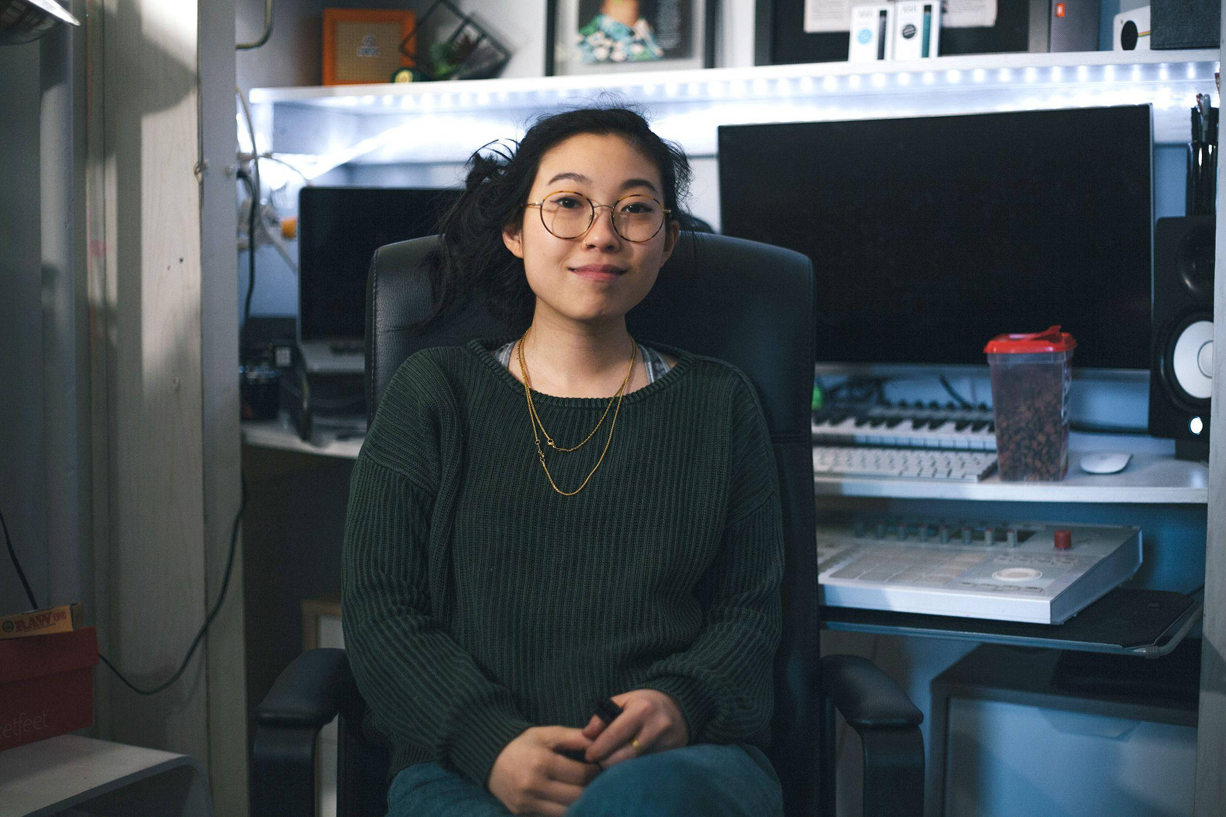 Awkwafina 桌布 壁紙 — 一位戴眼鏡的年輕女性微笑著坐在黑色辦公椅上，身處充滿科技感的家庭工作室，沐浴在柔和的藍色LED燈光中。