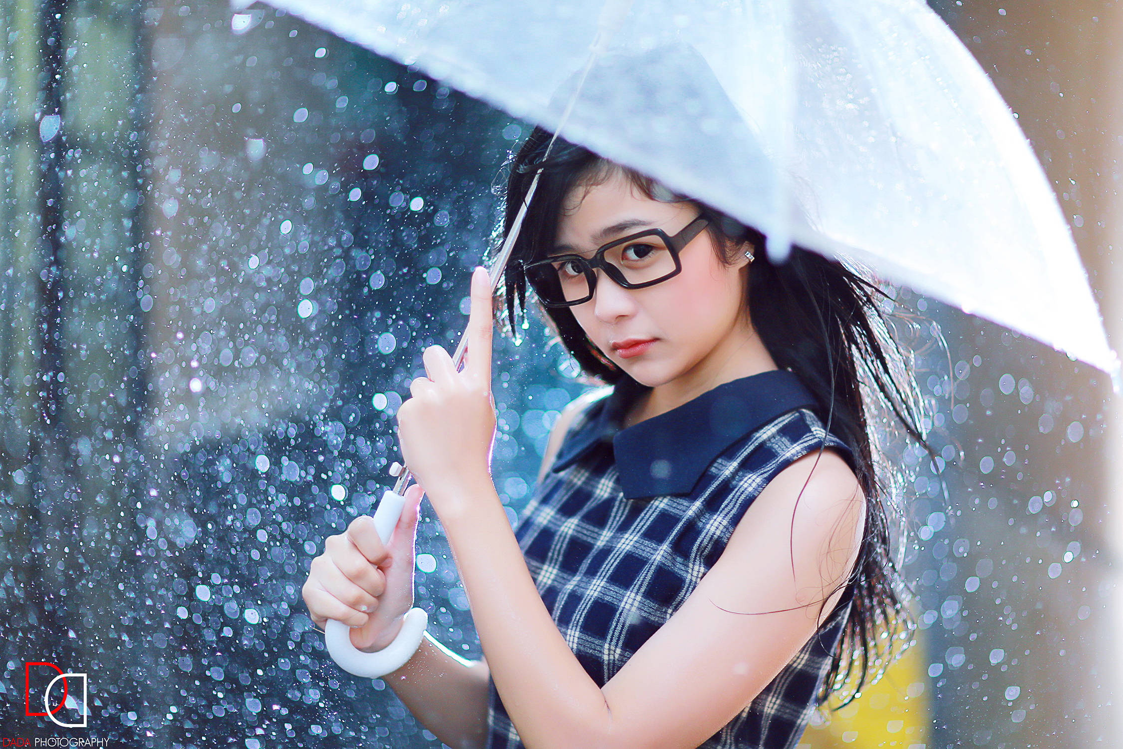 模特兒 桌布 壁紙 — 一位戴眼鏡的年輕女性拿著透明雨傘，雨滴在她周圍閃爍，營造出柔焦的城市場景。