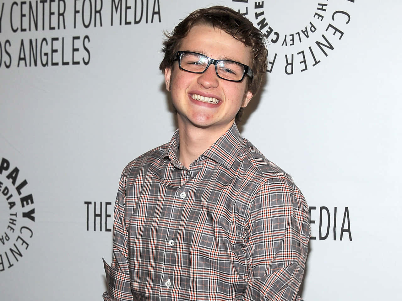 actor 桌布 壁紙 — 一位戴眼鏡的年輕男子在品牌活動背景前穿著格子襯衫，笑容燦爛。