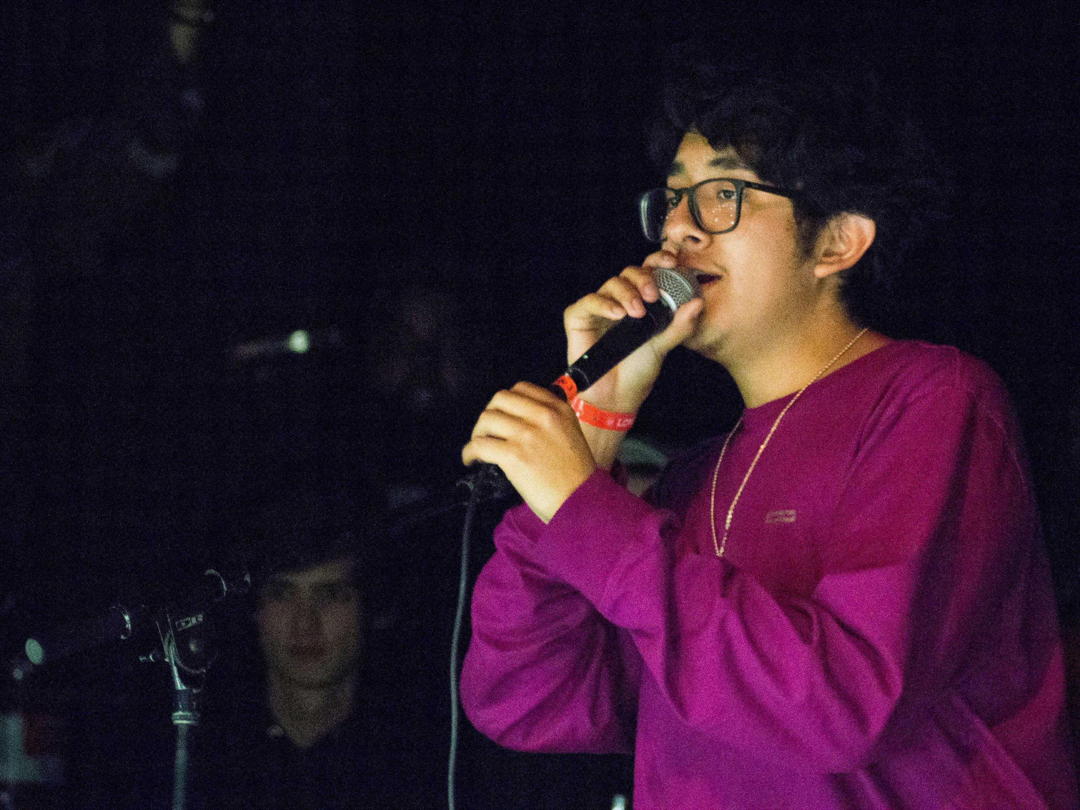 Singing 桌布 壁紙 — 一位戴眼鏡的年輕男子在昏暗的舞台燈光下，穿著鮮豔的紫色襯衫，熱情地對著麥克風唱歌。