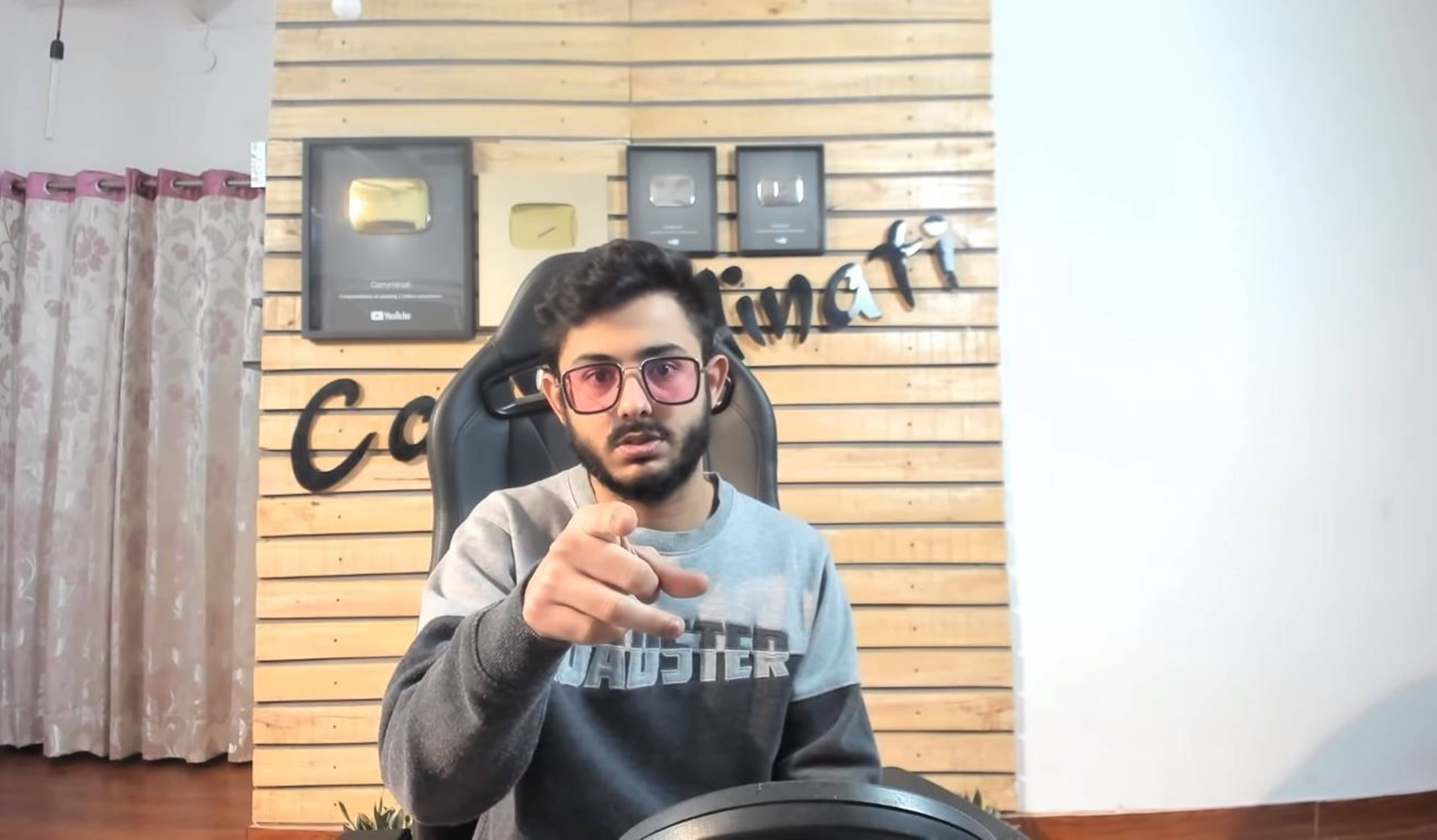 印度 桌布 壁紙 — 一位戴眼鏡的年輕男子坐在電競椅上，對著木牆上的YouTube播放按鈕，直接指向相機。