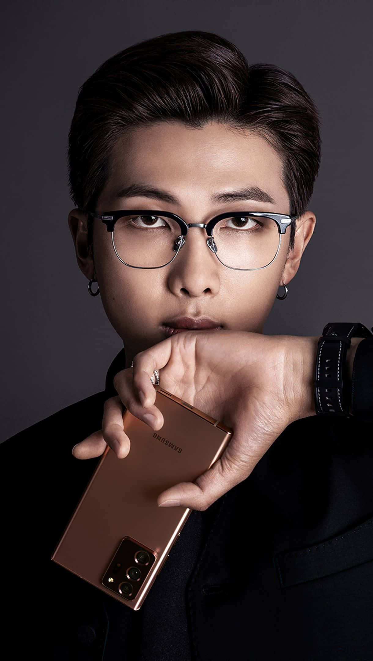 bts iPhone 桌布 壁紙 — 一位戴眼鏡的年輕男子手持玫瑰金色三星手機，在深灰色背景前凝視著。