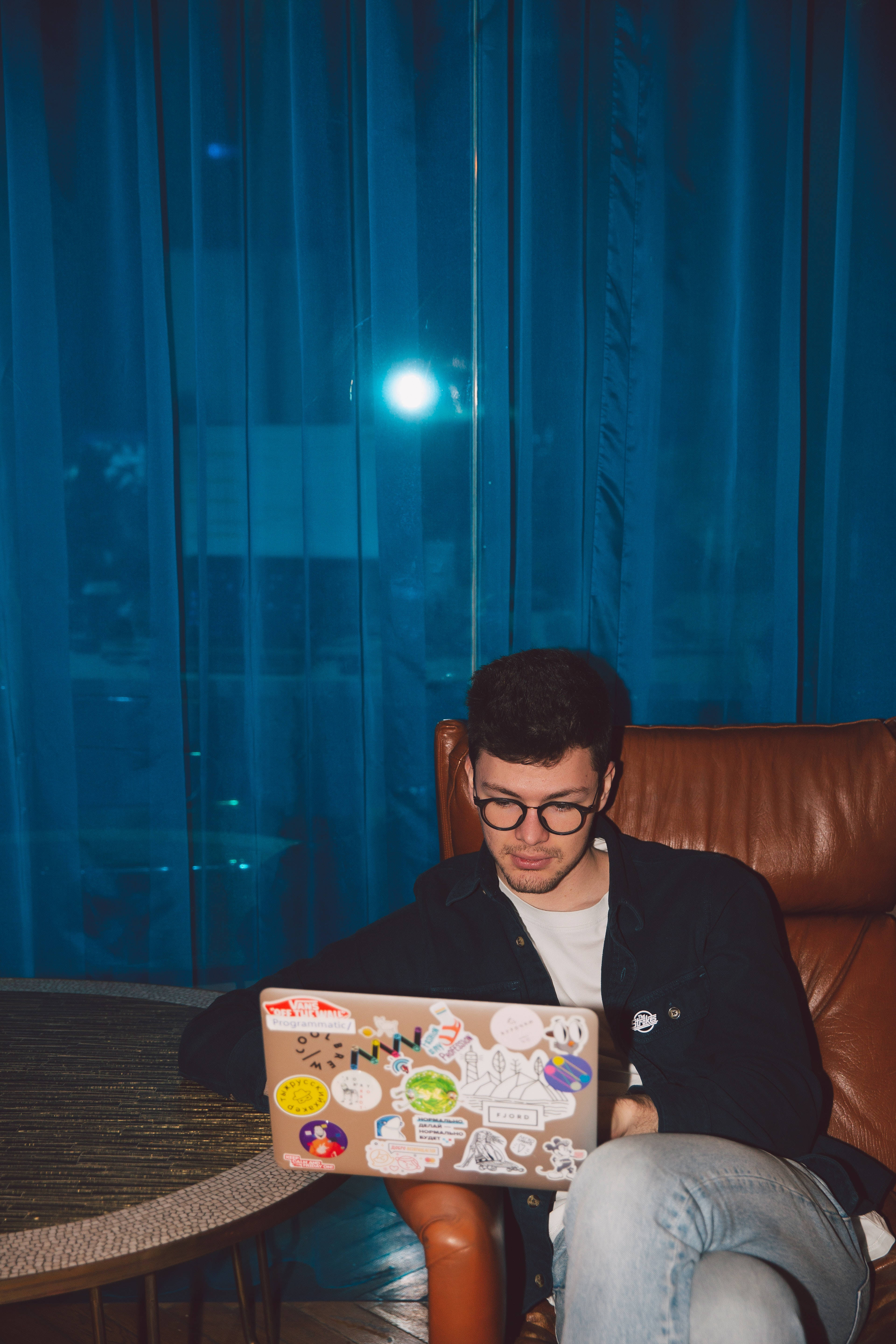 Cute Laptop 桌布 壁紙 — 一位戴眼鏡的年輕男子晚上坐在棕色皮椅上，專注於一台貼滿彩色貼紙的筆記型電腦，背景是藍色窗簾和明亮的窗光。