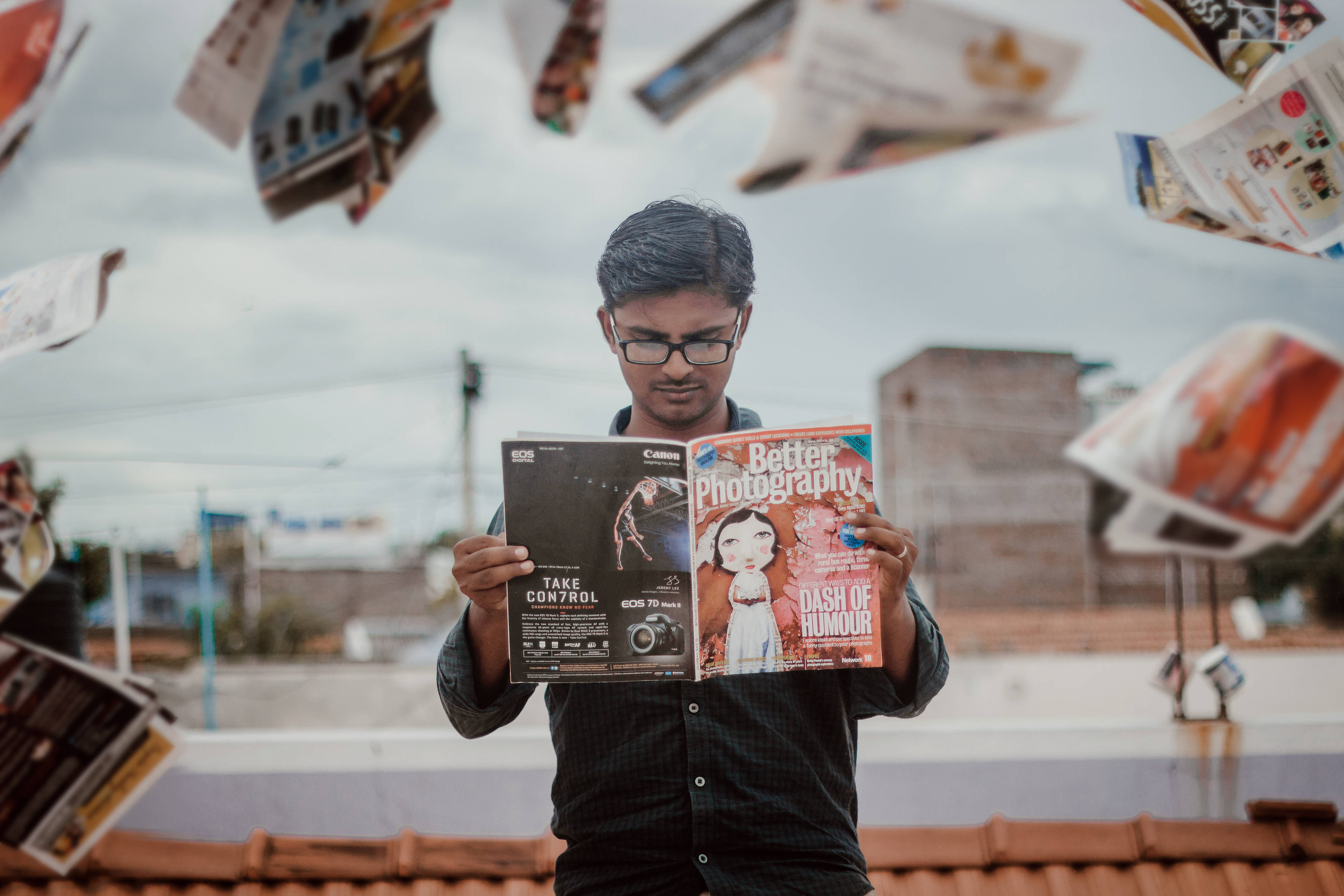 Magazine 桌布 壁紙 — 一位戴眼鏡的年輕男子正在閱讀兩本攝影雜誌，報紙在他周圍漂浮，構成超現實的城市場景。