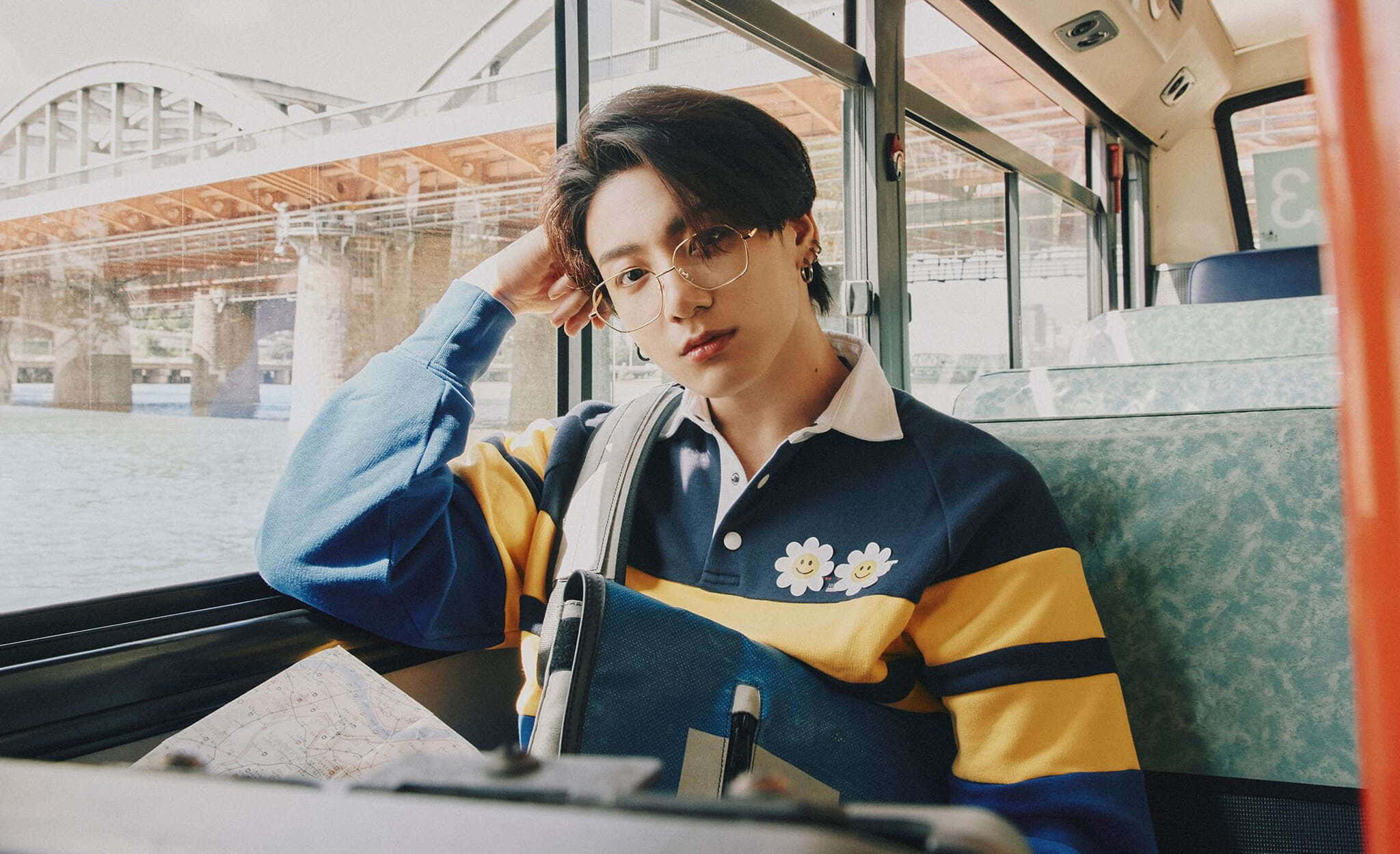 Jungkook 桌布 壁紙 — 一位戴眼鏡的年輕男子靠在巴士窗邊，俯瞰橋樑和水面，身穿藍黃條紋毛衣，帶有花朵補丁。