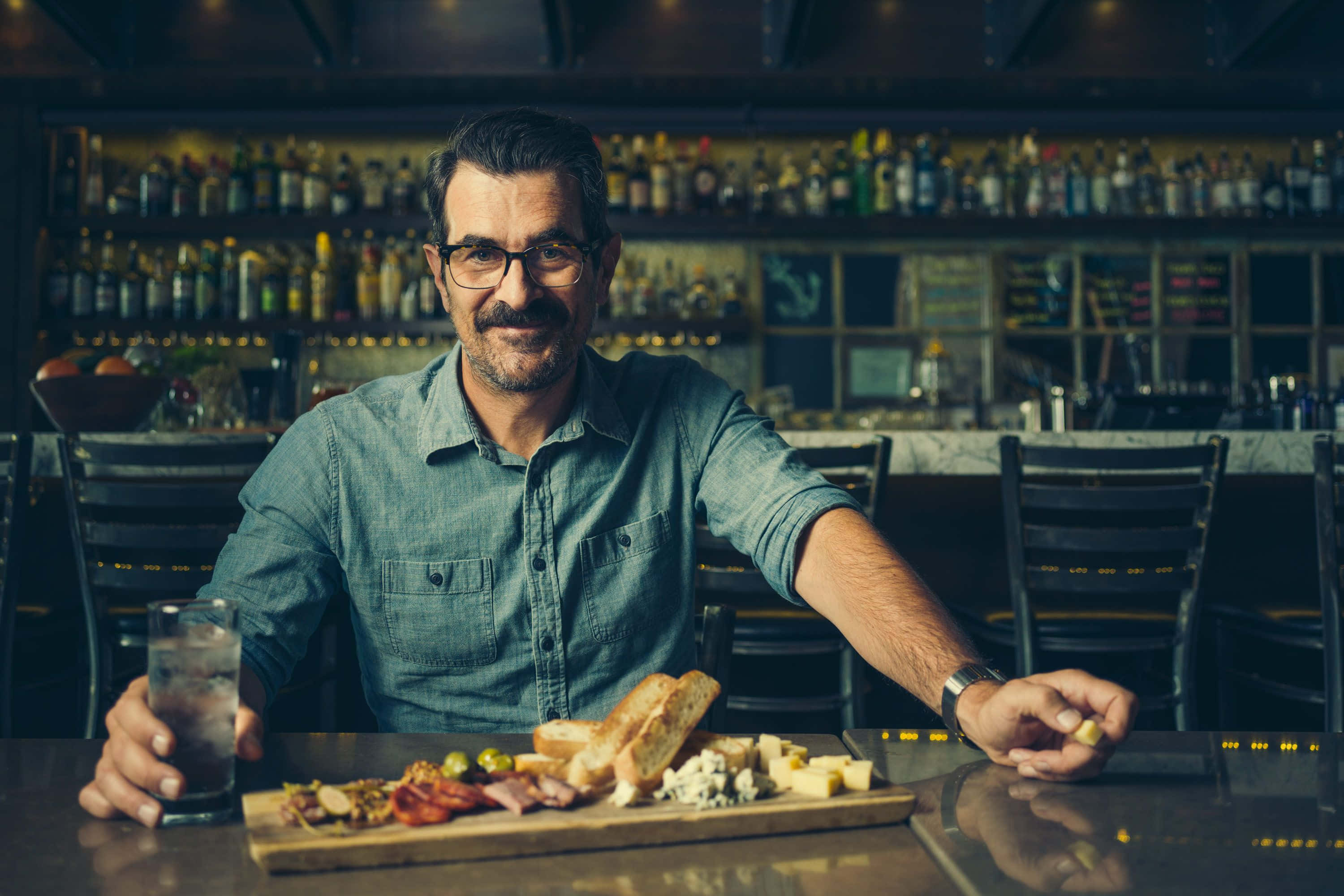 Ty Burrell 桌布 壁紙 — 一位戴眼鏡的微笑男士在燈光昏暗的酒吧裡，身後是擺滿酒瓶的架子，享受著熟食拼盤和飲料。
