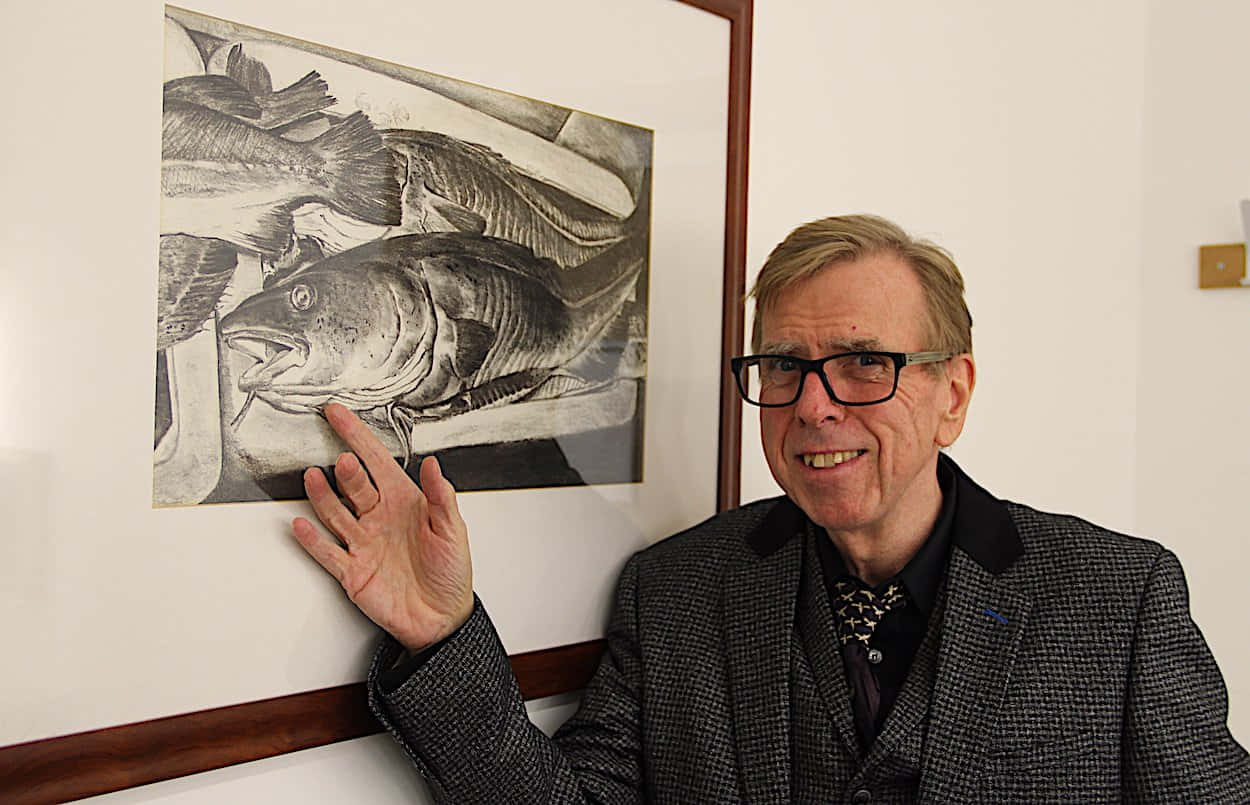 Timothy Spall 桌布 壁紙 — 一位戴眼鏡的微笑男士正指向白色牆上一個裝裱好的詳細鉛筆魚類畫作。