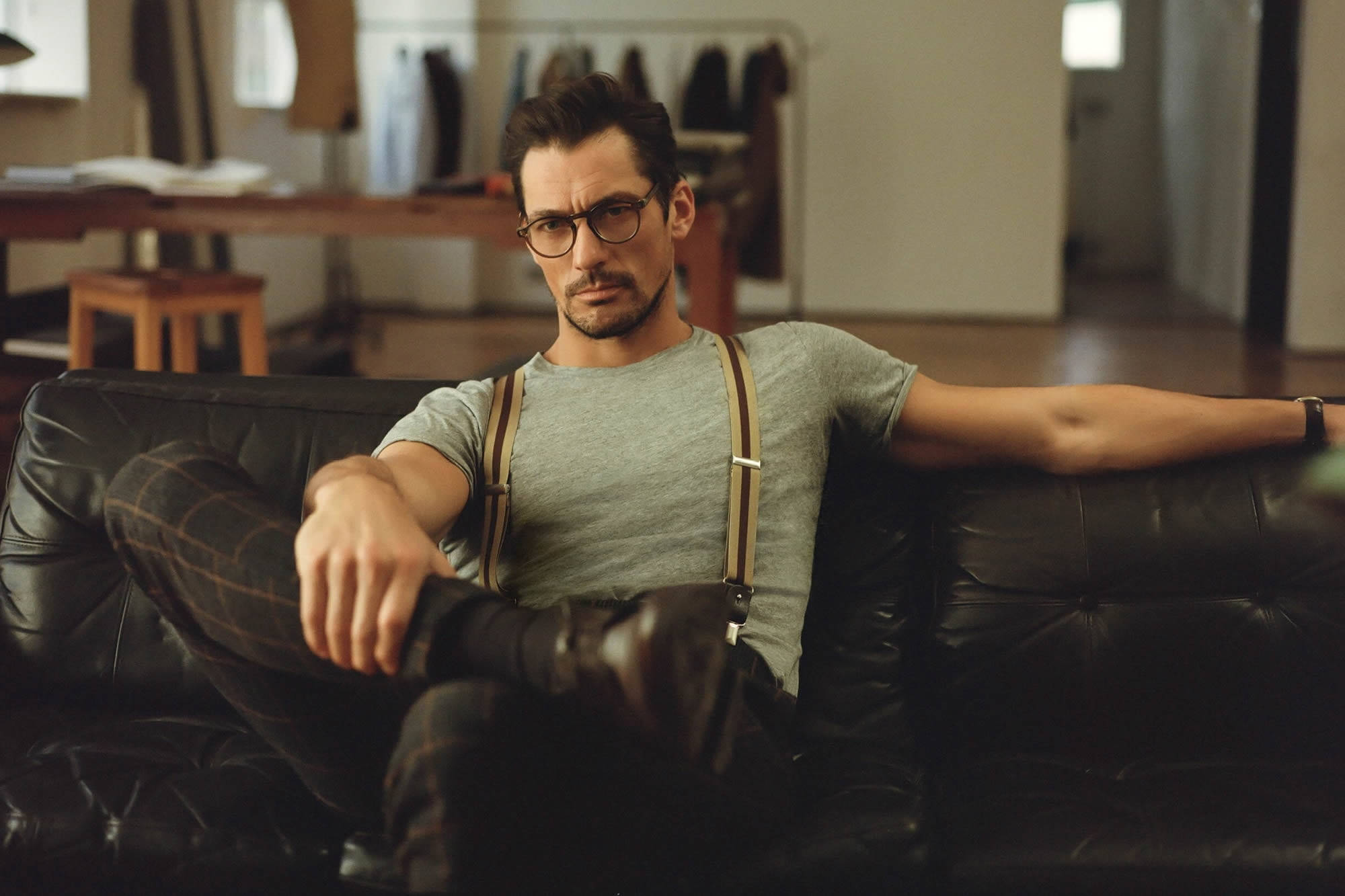 sexy man 桌布 壁紙 — 一位戴眼鏡的時尚男士自信地躺在一張黑色皮革沙發上，身處一個溫暖光線、簡約風格的室內空間。