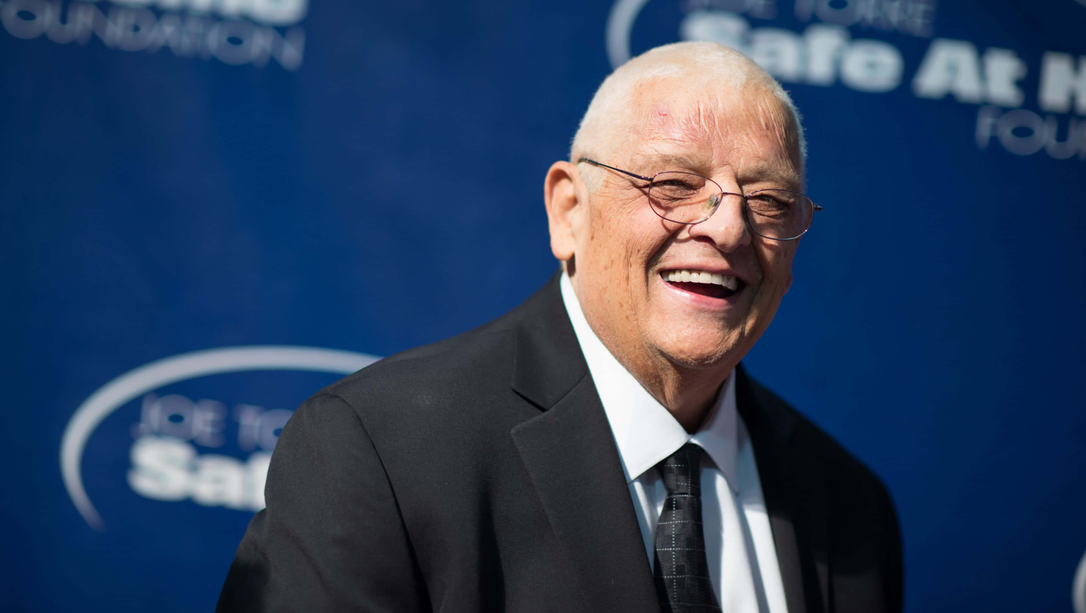 Dusty Rhodes 桌布 壁紙 — 一位戴眼鏡的較年長男士穿著黑色西裝，在藍色活動背景前開懷大笑。