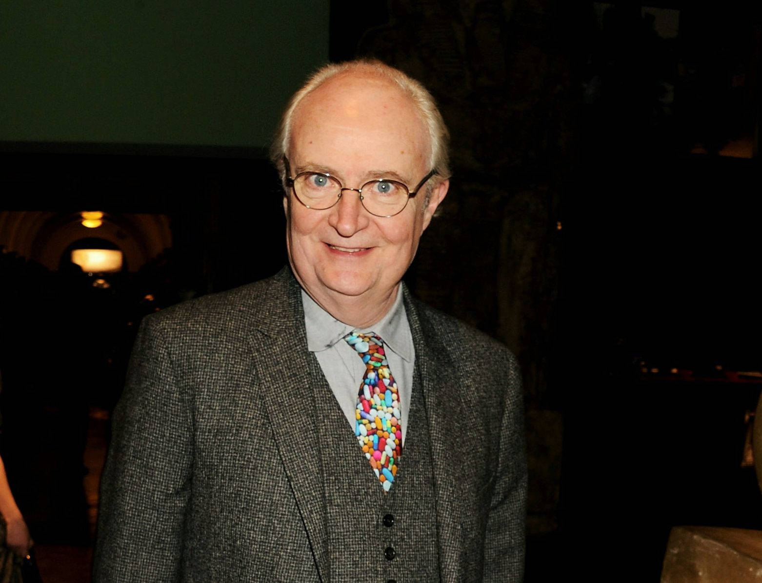 English 桌布 壁紙 — 一位戴眼鏡的長者，穿著粗花呢西裝和色彩繽紛的糖果圖案領帶，露出溫暖的笑容。