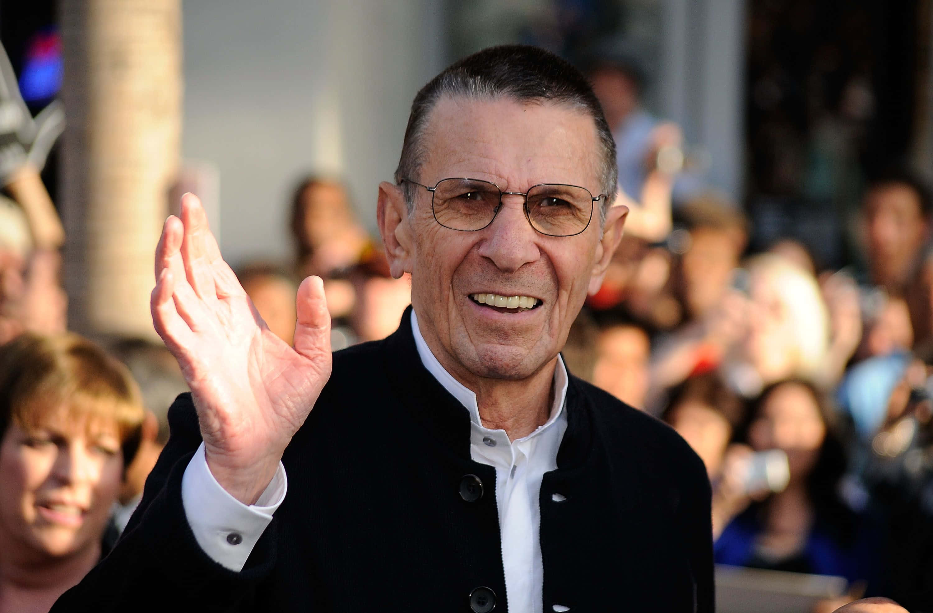 Celebrity 桌布 壁紙 — 一位戴眼鏡的長者在陽光明媚的戶外活動中愉快地向人群揮手。