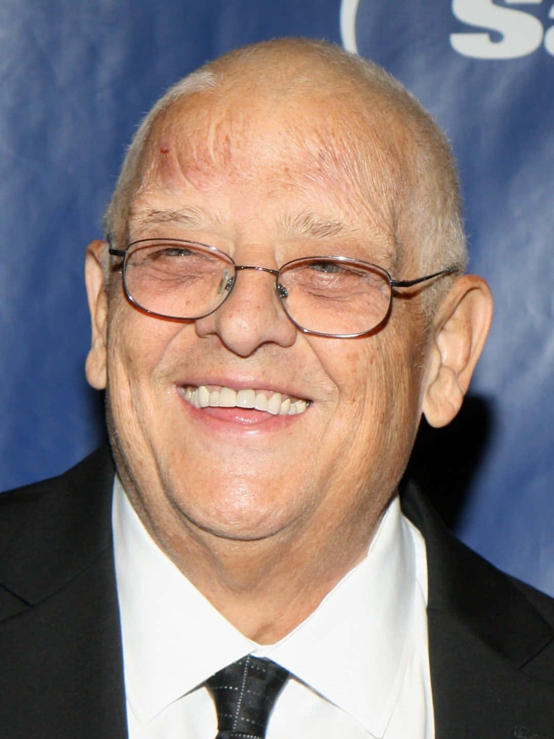 Dusty Rhodes 桌布 壁紙 — 一位戴眼鏡的長者身穿黑色西裝，在藍色背景下溫暖地微笑。