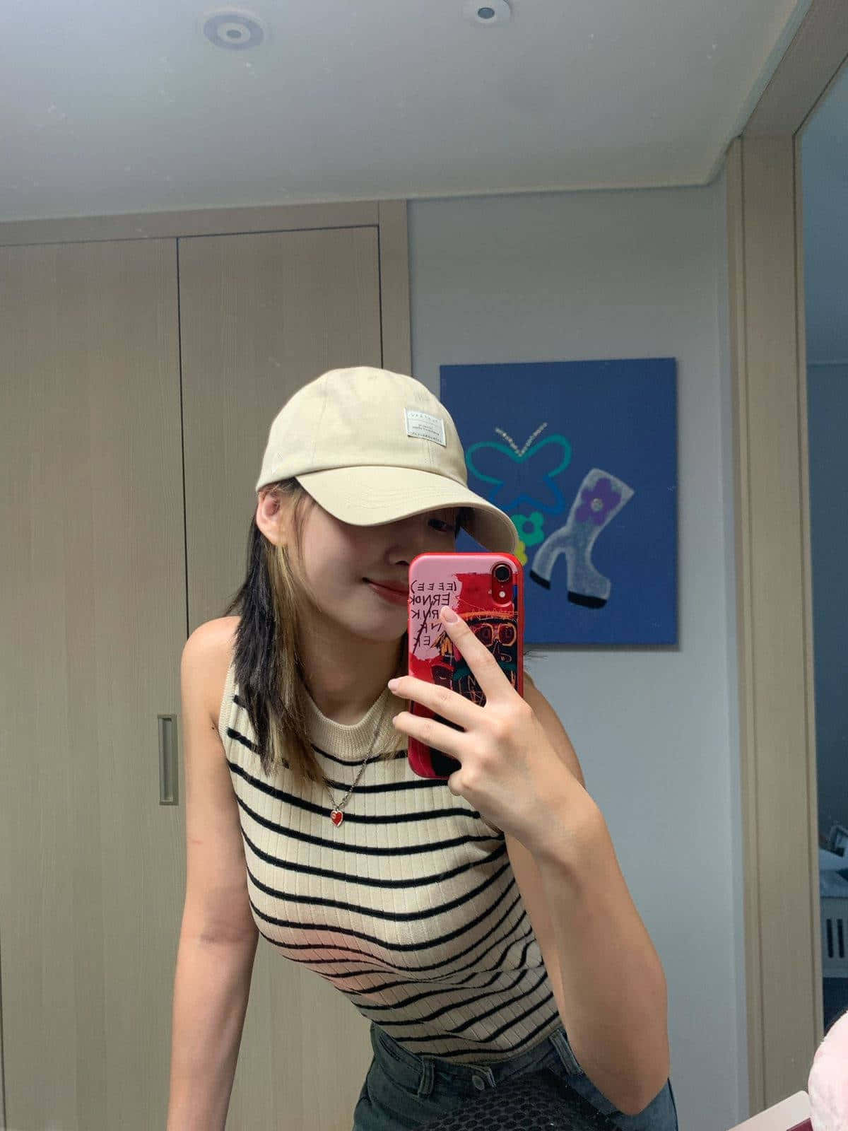 美學 桌布 壁紙 — 一位戴米色帽子和條紋上衣的年輕女性在現代房間裡用紅色手機對著鏡子自拍。