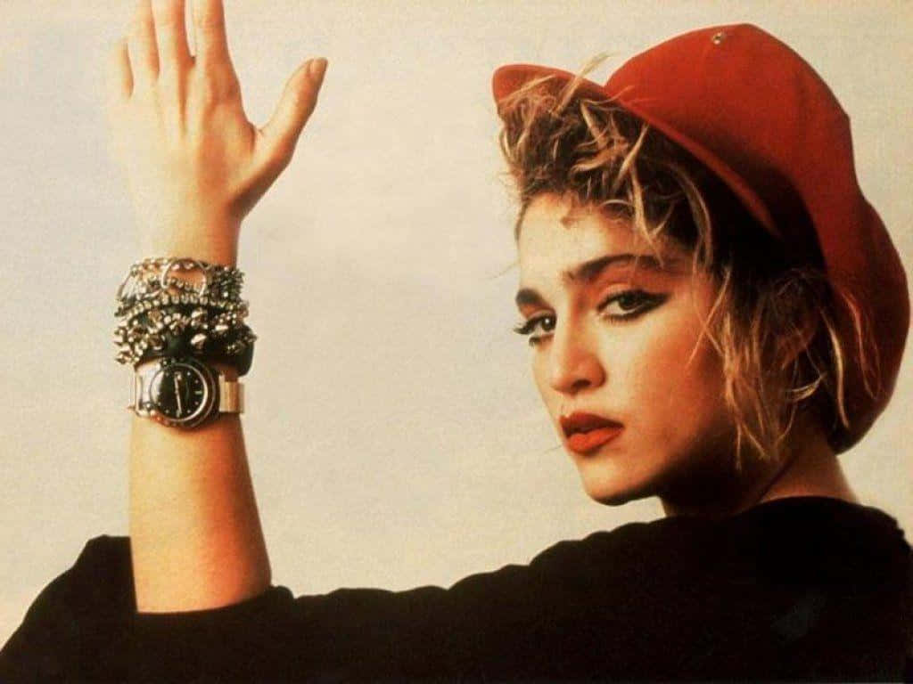 Madonna 桌布 壁紙 — 一位戴紅帽、穿黑色上衣的女性，在純色背景前舉起戴有層疊手鐲和手錶的手。