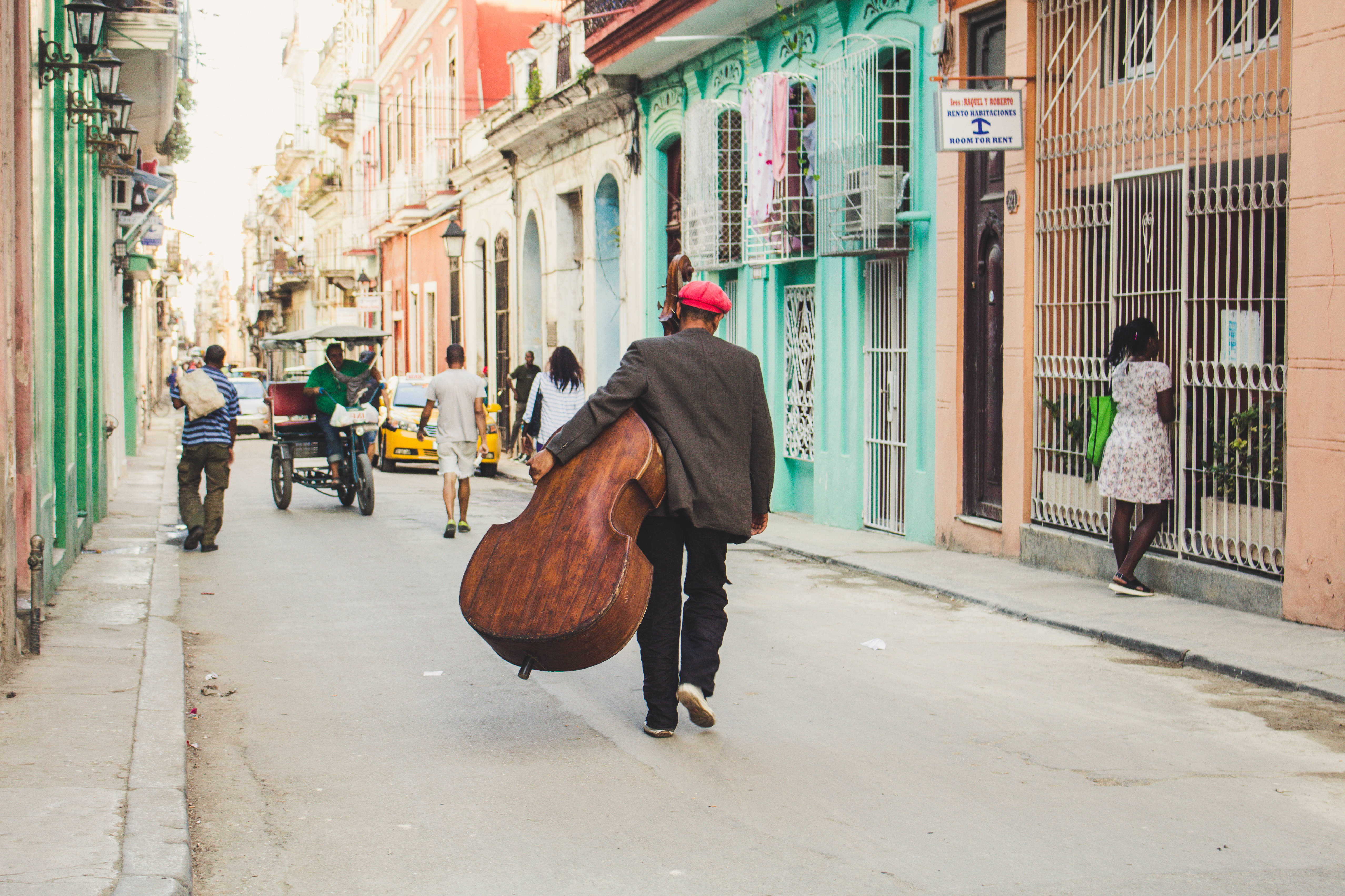 Cuba 桌布 壁紙 — 一位戴紅帽的音樂家背著一個大提琴，走在一條充滿活力、狹窄的街道上，兩旁是色彩繽紛的殖民時期建築。