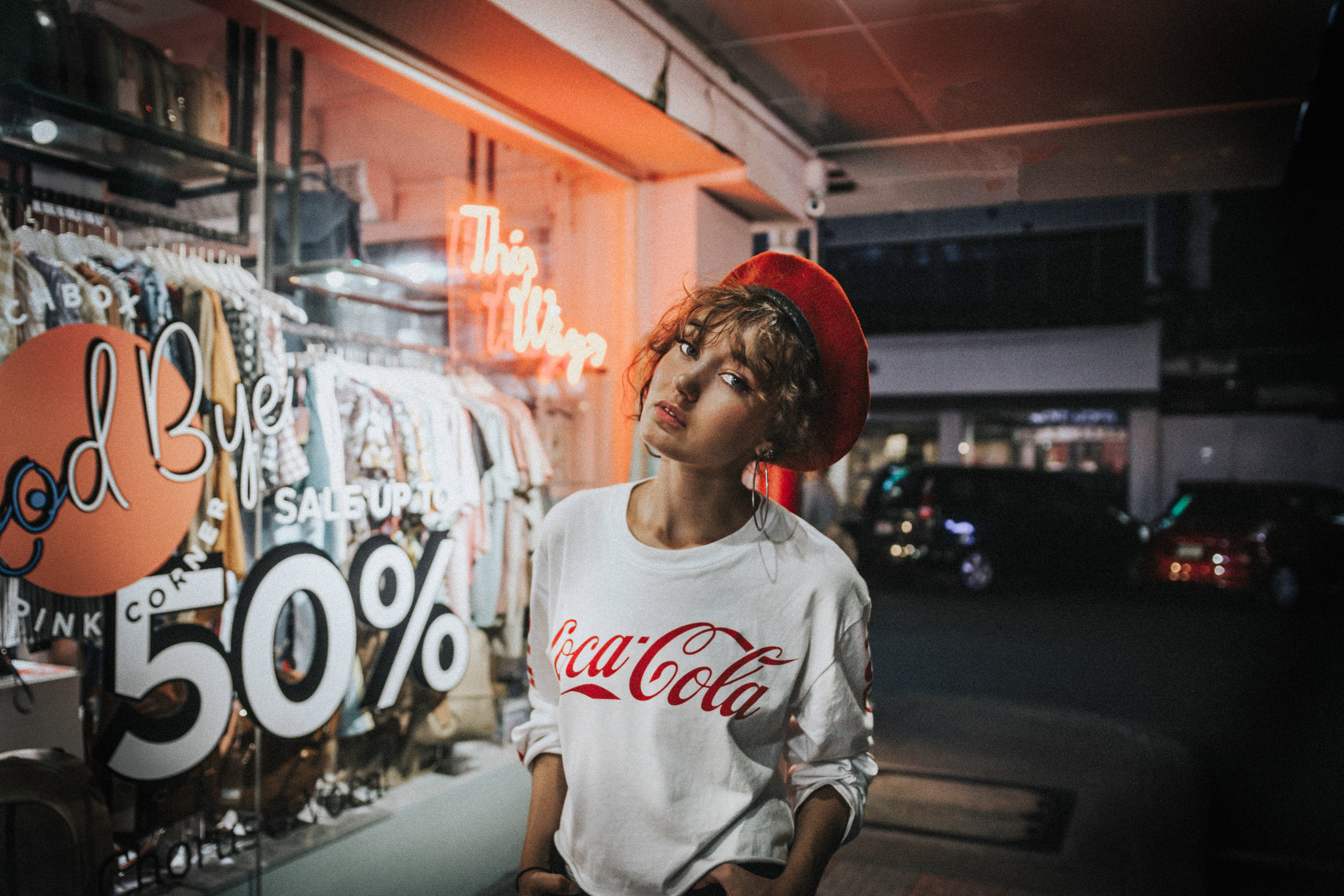Trendy 桌布 壁紙 — 一位戴紅色貝雷帽、身穿可口可樂T恤的年輕女子夜晚站在霓虹燈照亮的商店櫥窗前。