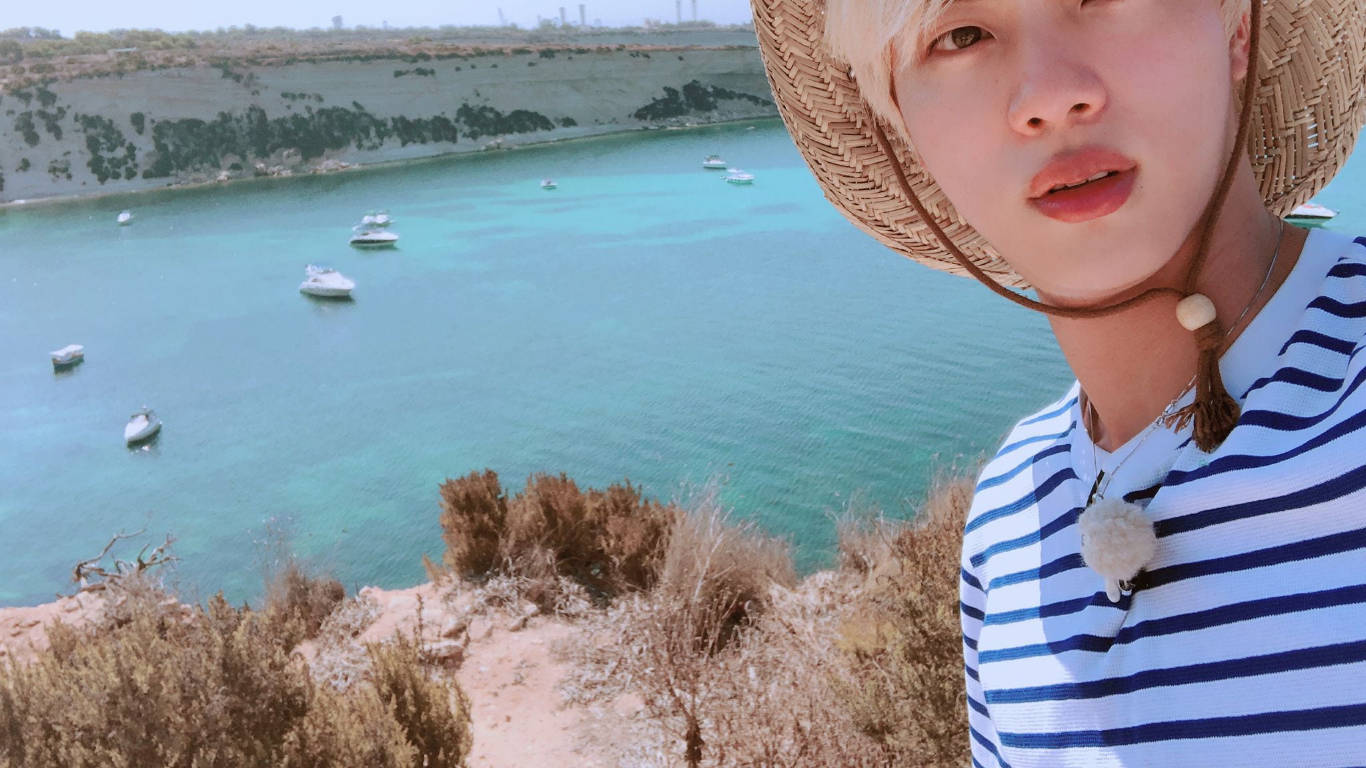 kim seok jin 桌布 壁紙 — 一位戴草帽、穿條紋襯衫的年輕人，以點綴著船隻和乾燥懸崖的碧綠海水為背景擺姿勢。