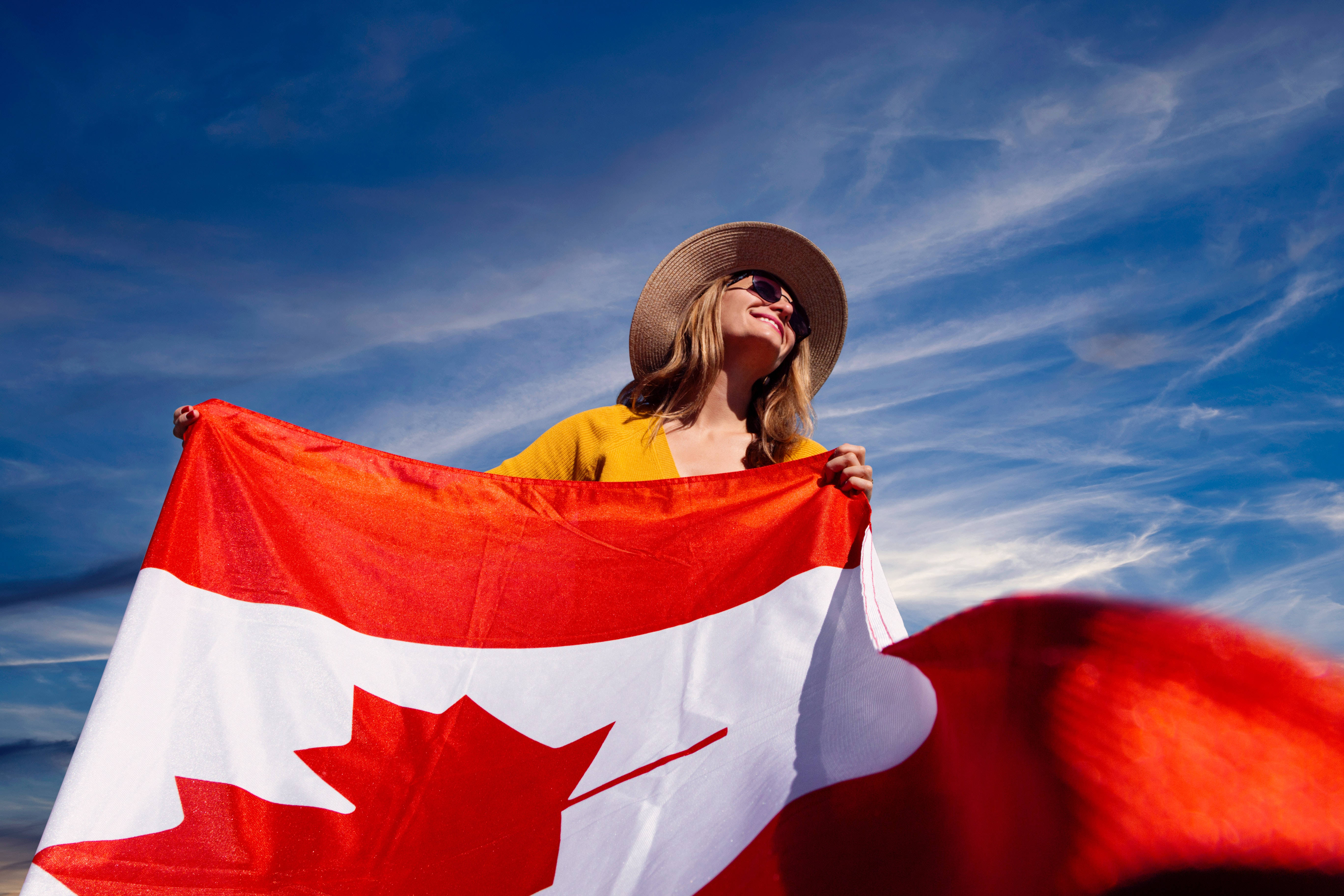 Flag 桌布 壁紙 — 一位戴草帽、穿黃色上衣的微笑女士，在藍天白雲下揮舞著加拿大國旗。