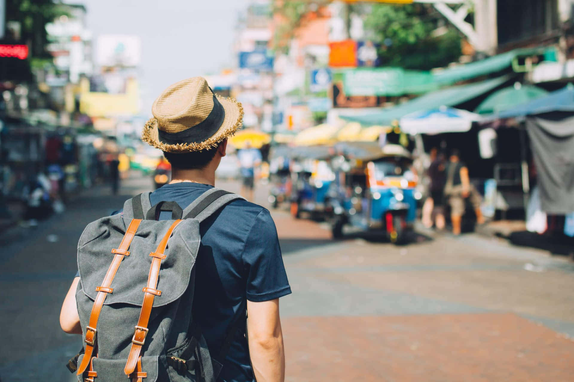 Travel Desktop 桌布 壁紙 — 一位戴草帽、背背包的旅客走在熱鬧的街道上，兩旁是色彩繽紛的市場攤位和嘟嘟車。