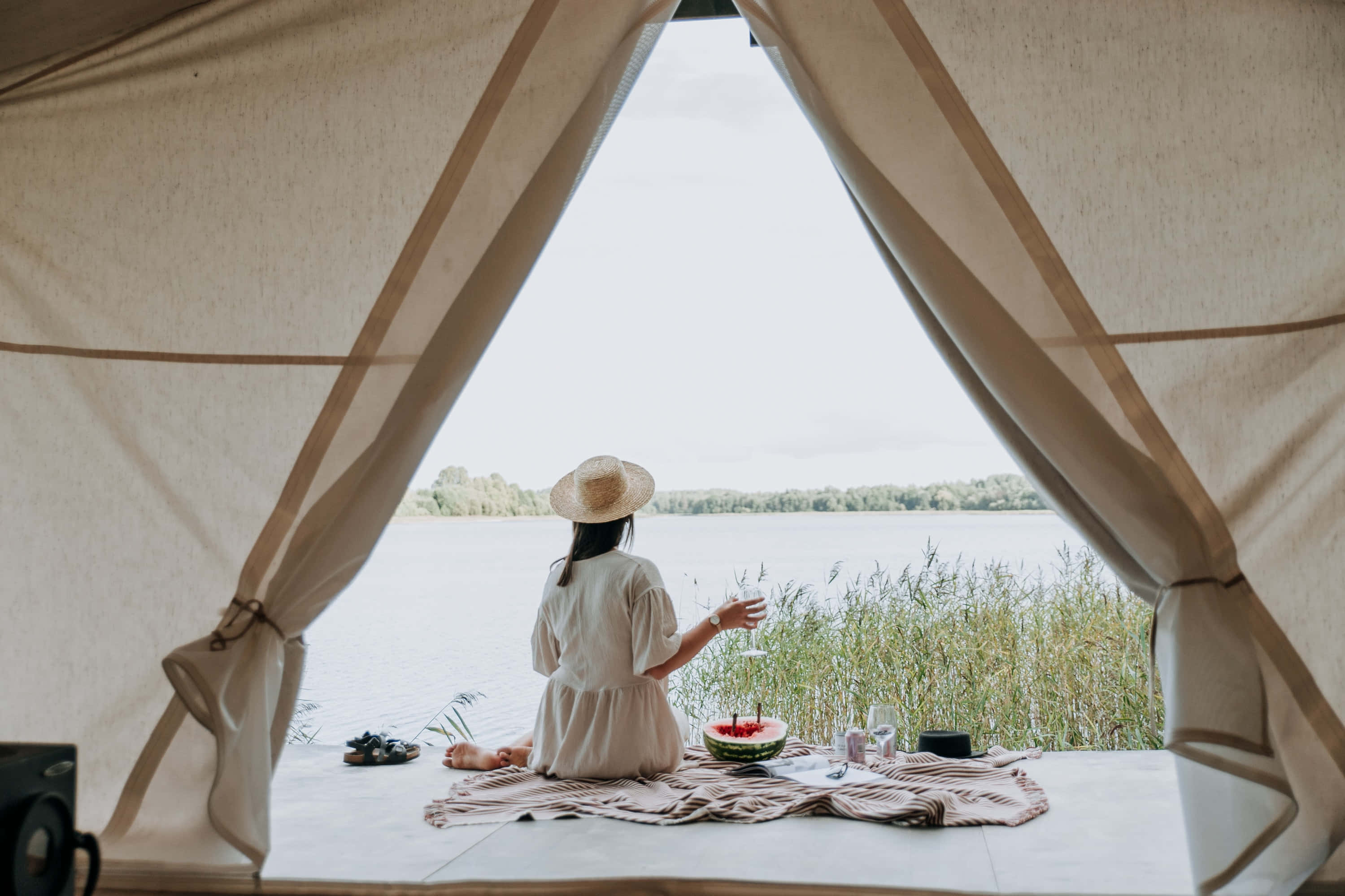 Camping 桌布 壁紙 — 一位戴草帽的女人坐在帆布帳篷內，在平靜的湖邊啜飲葡萄酒，湖邊有蘆葦和遠處的樹木，天空多雲。