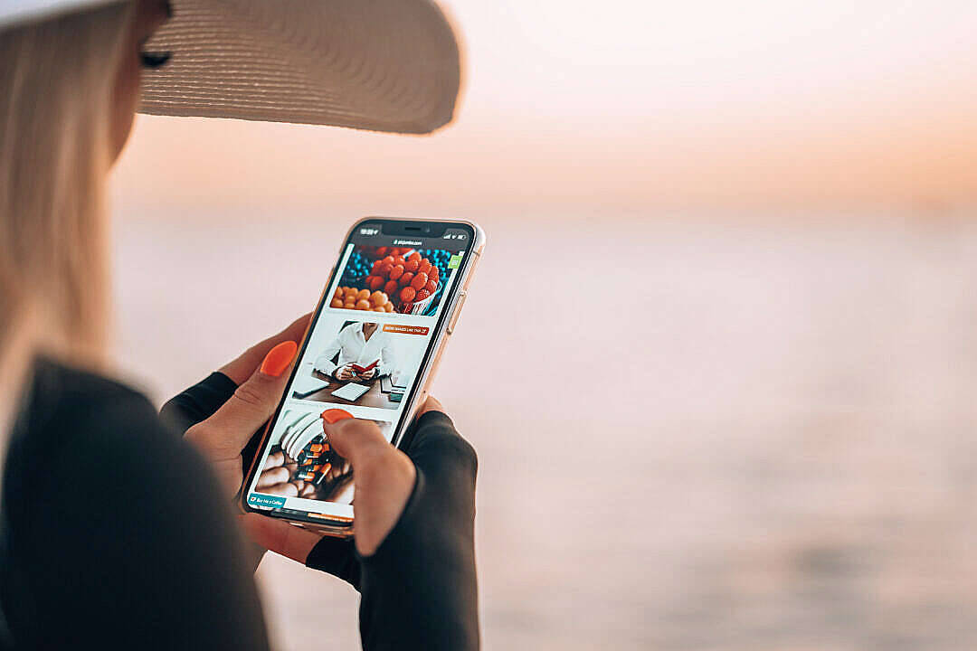 photography iPhone 桌布 壁紙 — 一位戴草帽的女士在寧靜的日落海濱用手機瀏覽食物照片。