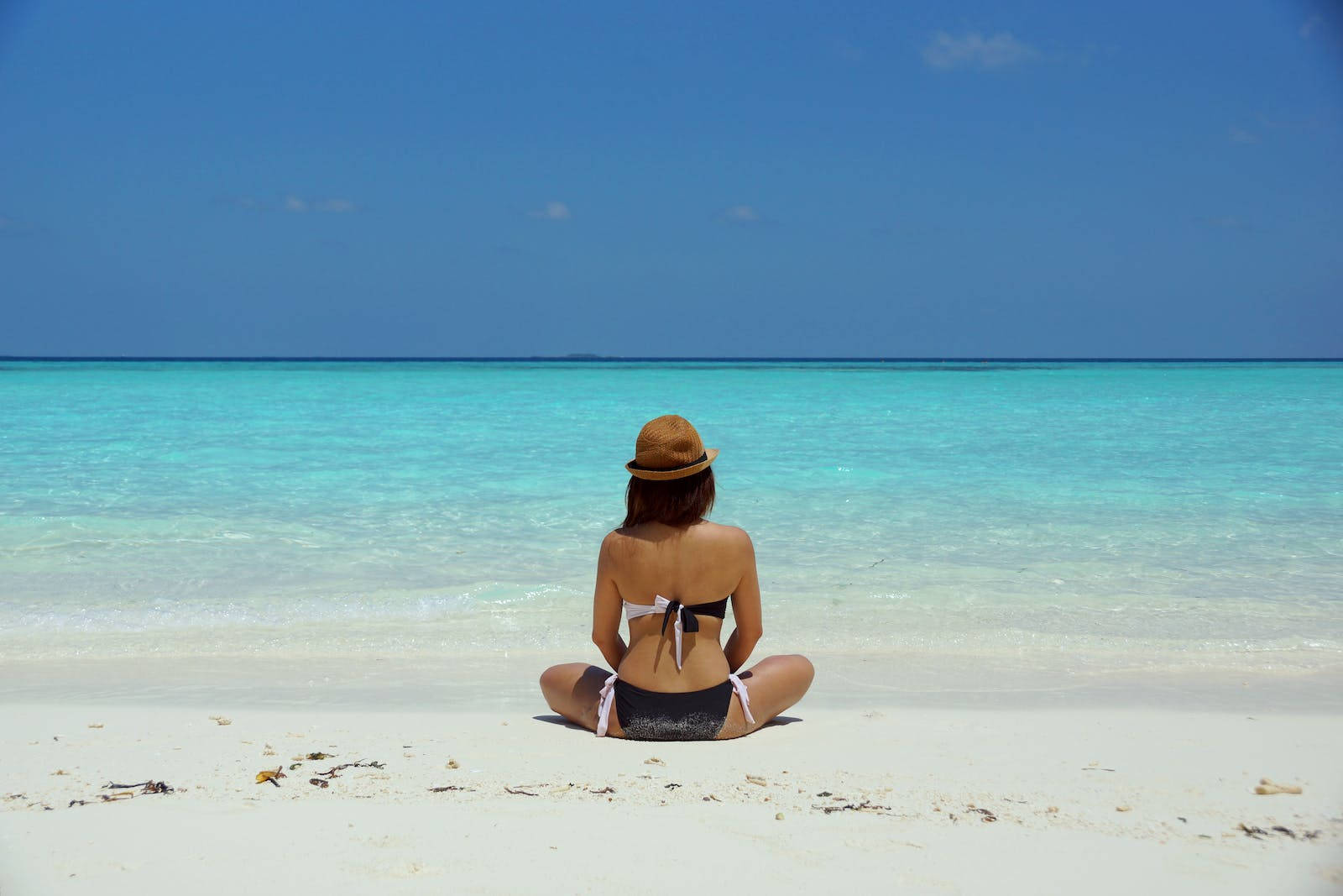 Relaxing 桌布 壁紙 — 一位戴草帽的女性在純淨的白色沙灘上盤腿而坐，面向湛藍清澈的天空下碧綠色的海水。