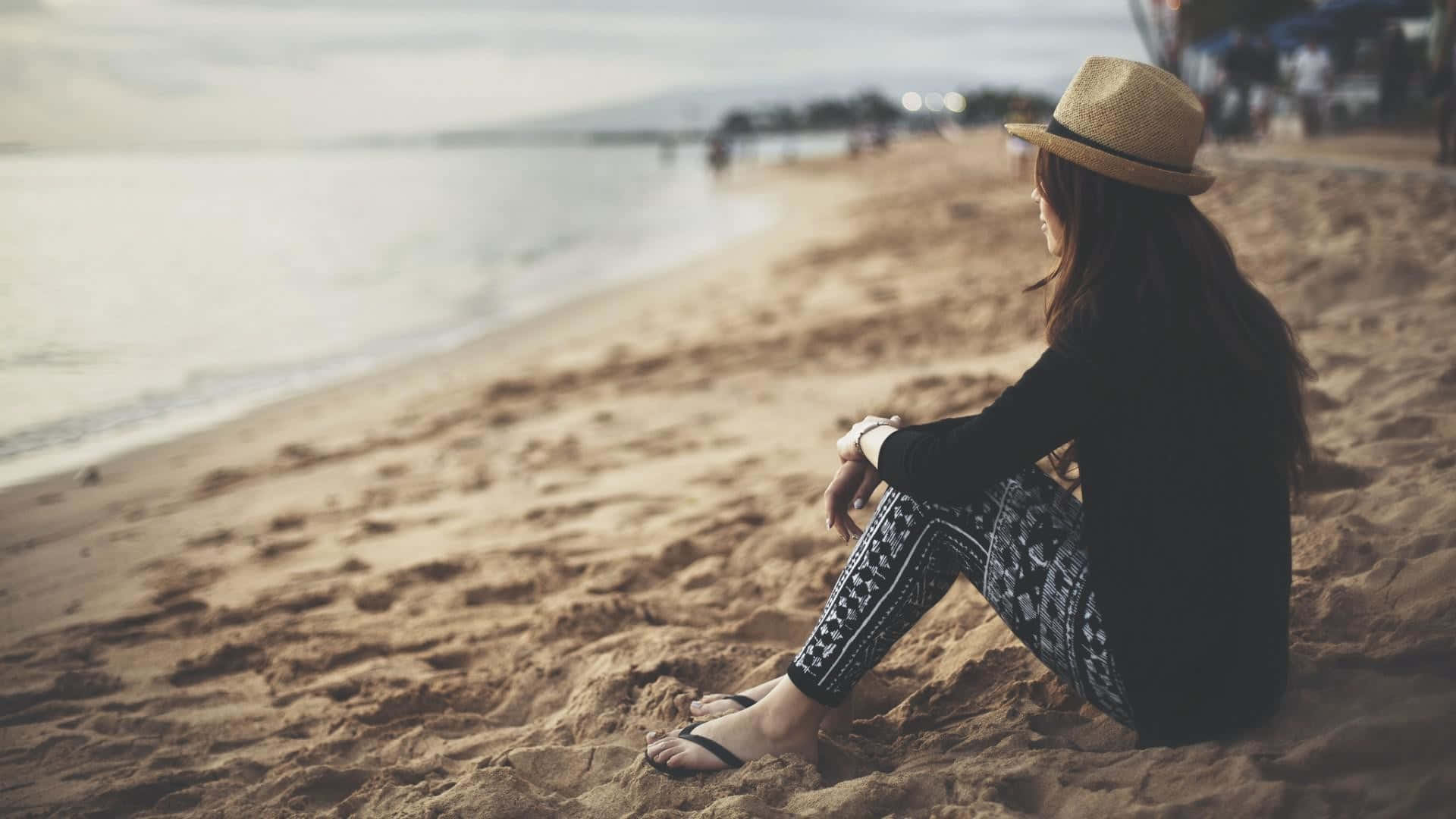 海馬 桌布 壁紙 — 一位戴草帽的女性獨自坐在沙灘上，凝視著柔和暮色下平靜的海洋。
