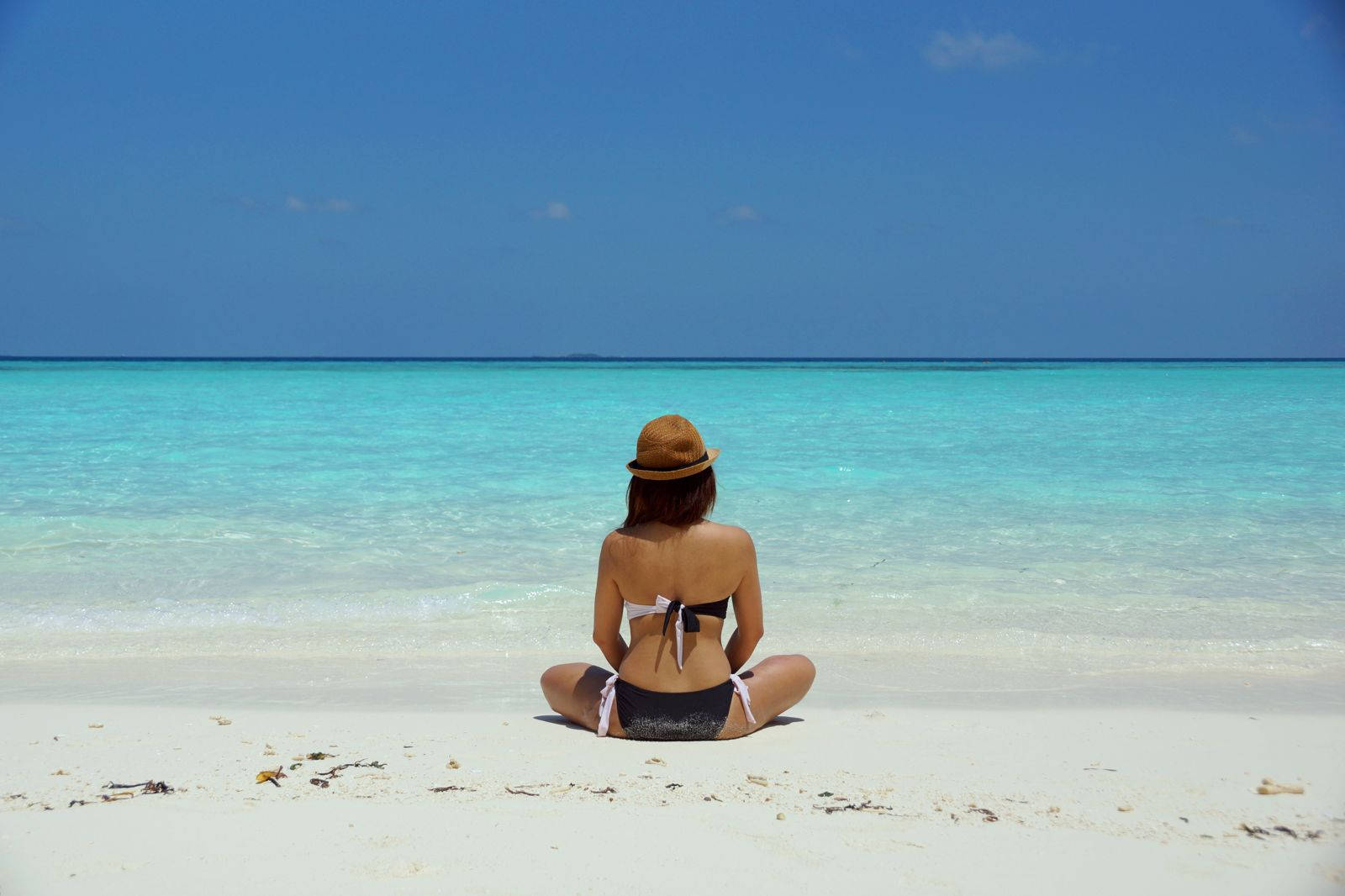Beach Girl 桌布 壁紙 — 一位戴草帽的女性盤腿坐在白色沙灘上，面向碧藍色的海洋，天空晴朗蔚藍。