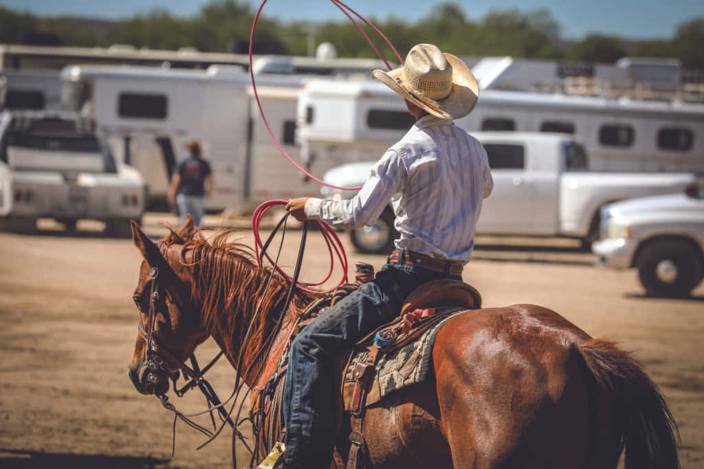 Cowboy 桌布 壁紙 — 一位戴草帽的牛仔騎著一匹棕馬，在塵土飛揚的牛仔競技場上揮舞紅色套索，背景有露營車和卡車。