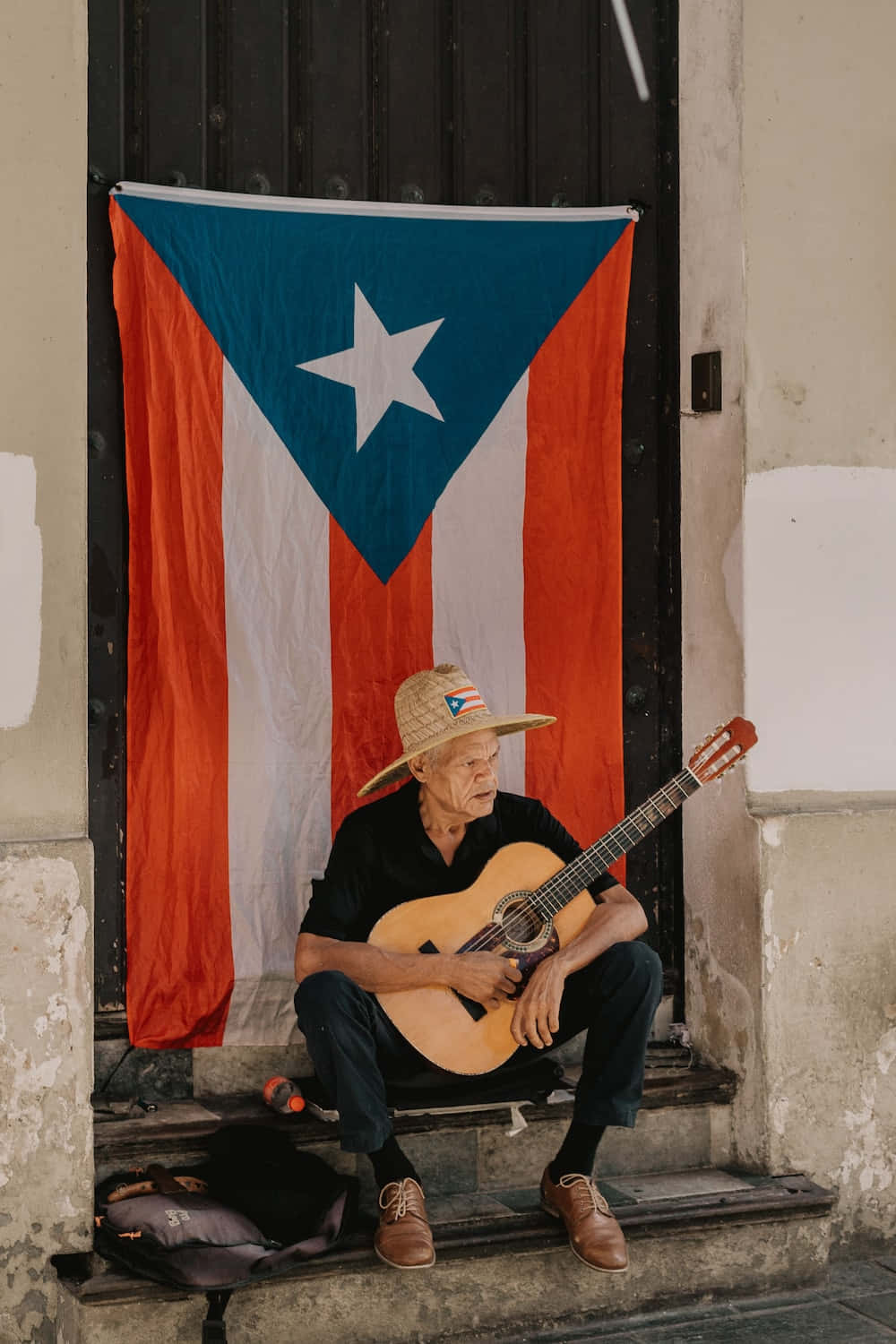 Puerto Rican 桌布 壁紙 — 一位戴草帽的長者坐在風化的台階上，在掛在門上的波多黎各國旗下彈吉他。