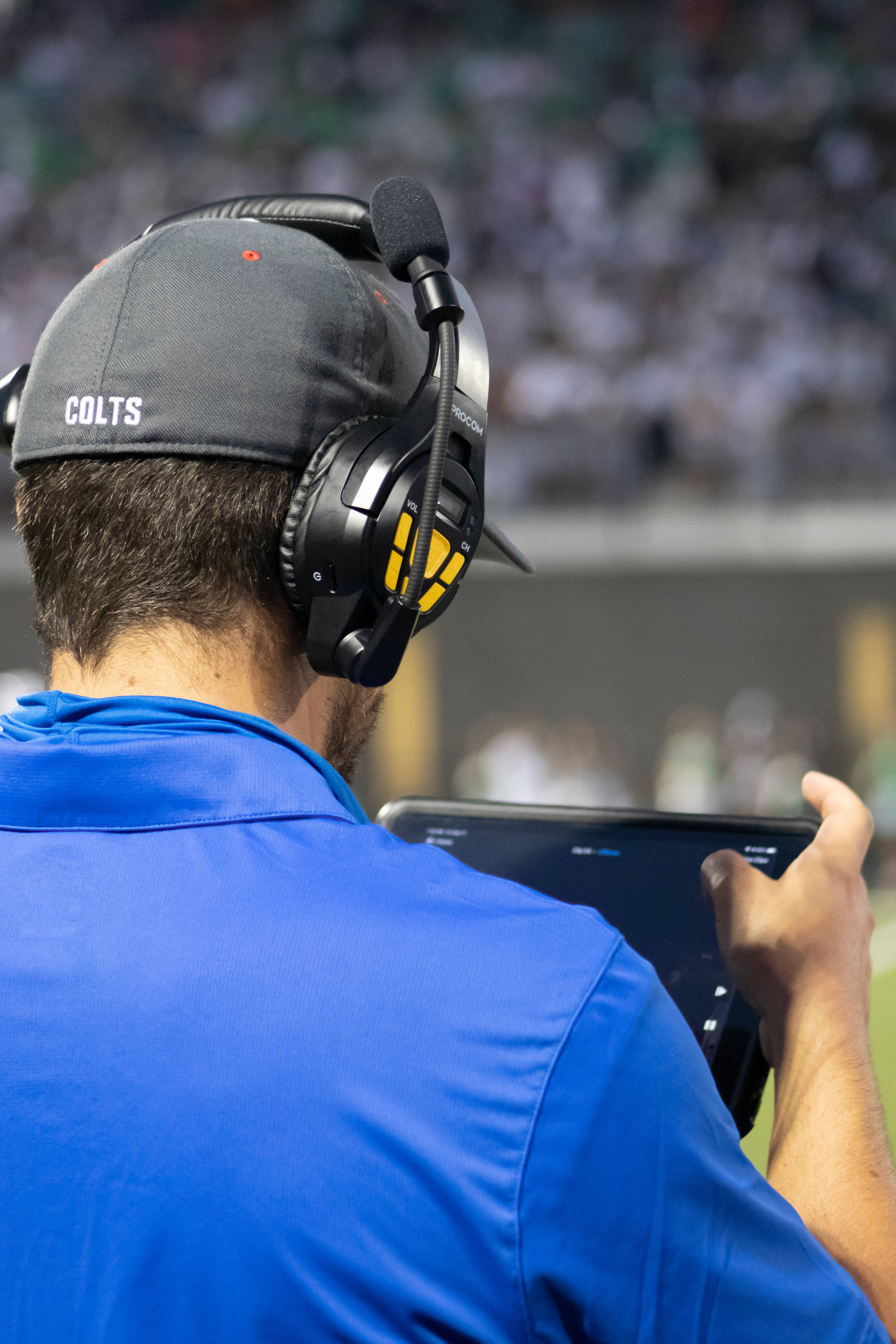NFL 美式足球 桌布 壁紙 — 一位戴著Colts帽和耳機的教練在足球比賽中專注地看著平板電腦，背景是模糊的體育場人群。