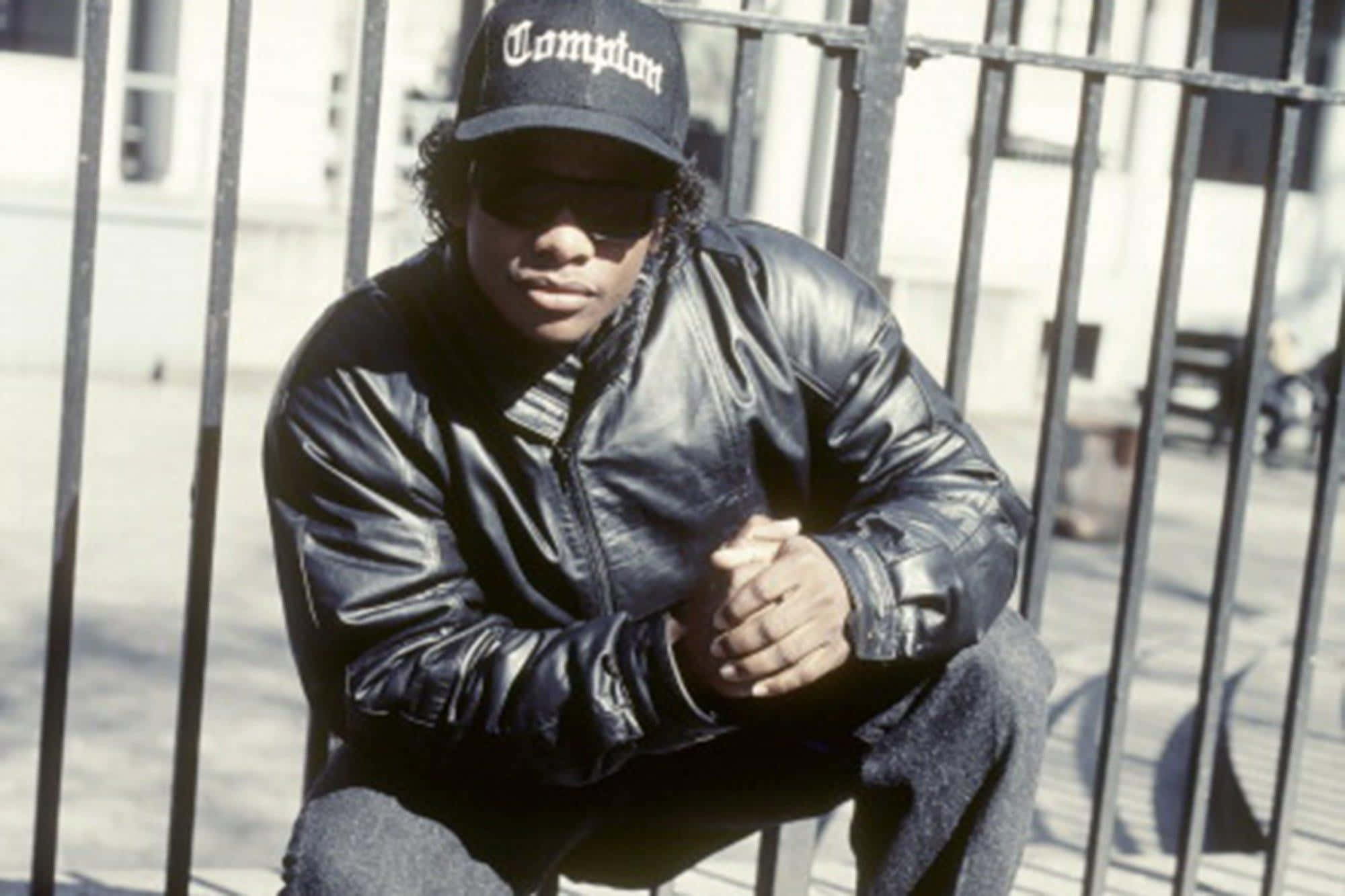 Eazy E 桌布 壁紙 — 一位戴著Compton帽和皮夾克的年輕男子自信地坐在金屬圍欄上，在明亮的日光下。