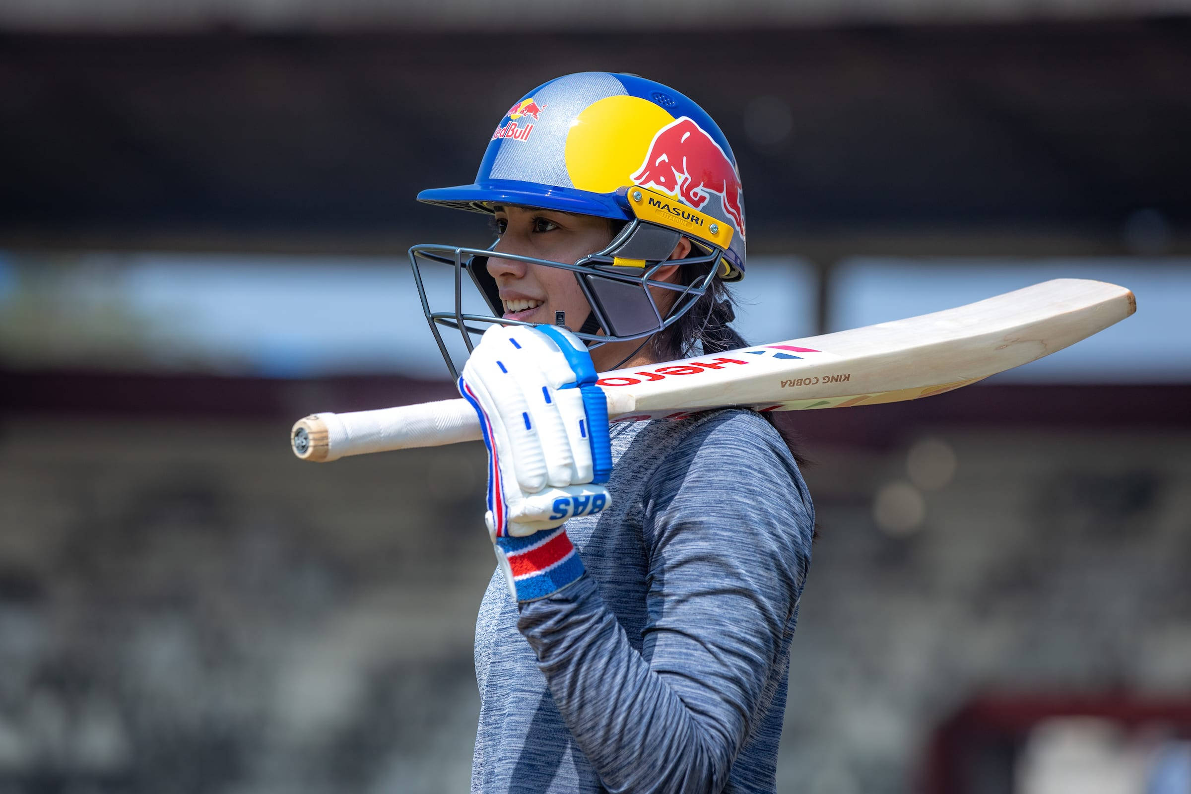 Smriti Mandhana 桌布 壁紙 — 一位戴著Red Bull頭盔、面帶微笑的女板球運動員，在陽光明媚的球場上將球棒扛在肩上。
