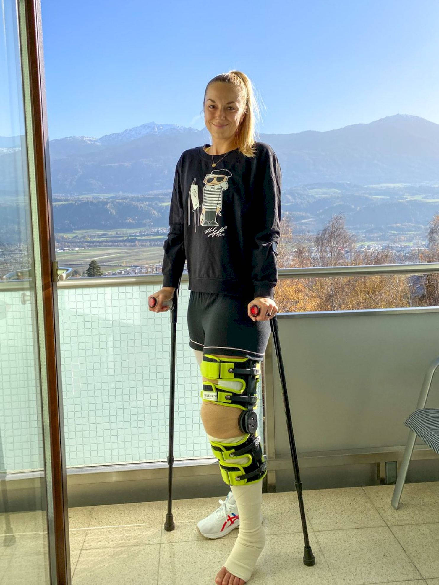 網球 桌布 壁紙 — 一位戴著亮綠色護膝和拐杖的女性在陽台上微笑，陽台可俯瞰白雪覆蓋的山脈，天空湛藍清澈。