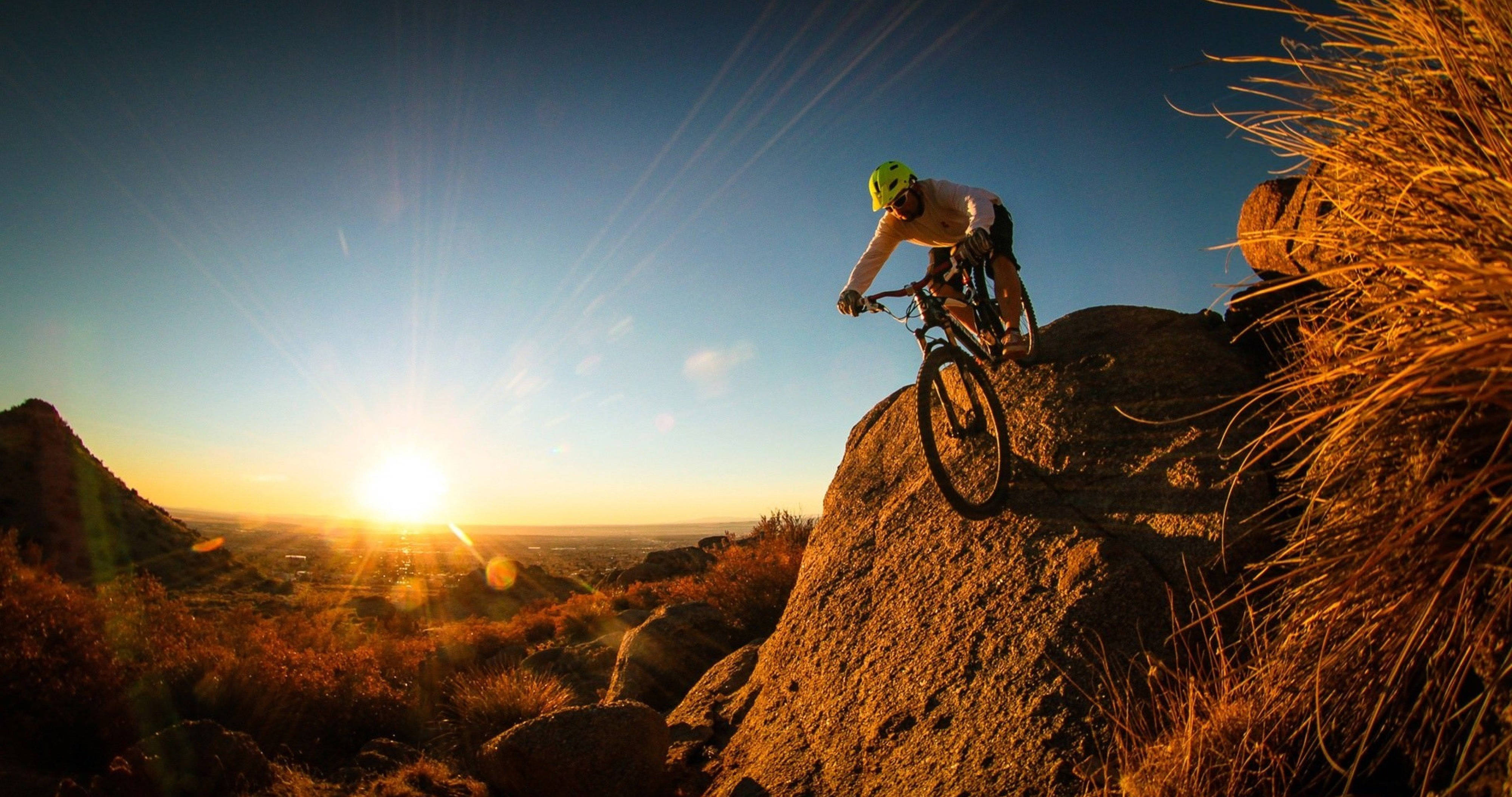 Mountain Bike 桌布 壁紙 — 一位戴著亮色頭盔的山地車手在岩石小徑上騎行，夕陽在他身後落下，將金色光芒灑在沙漠景觀上。