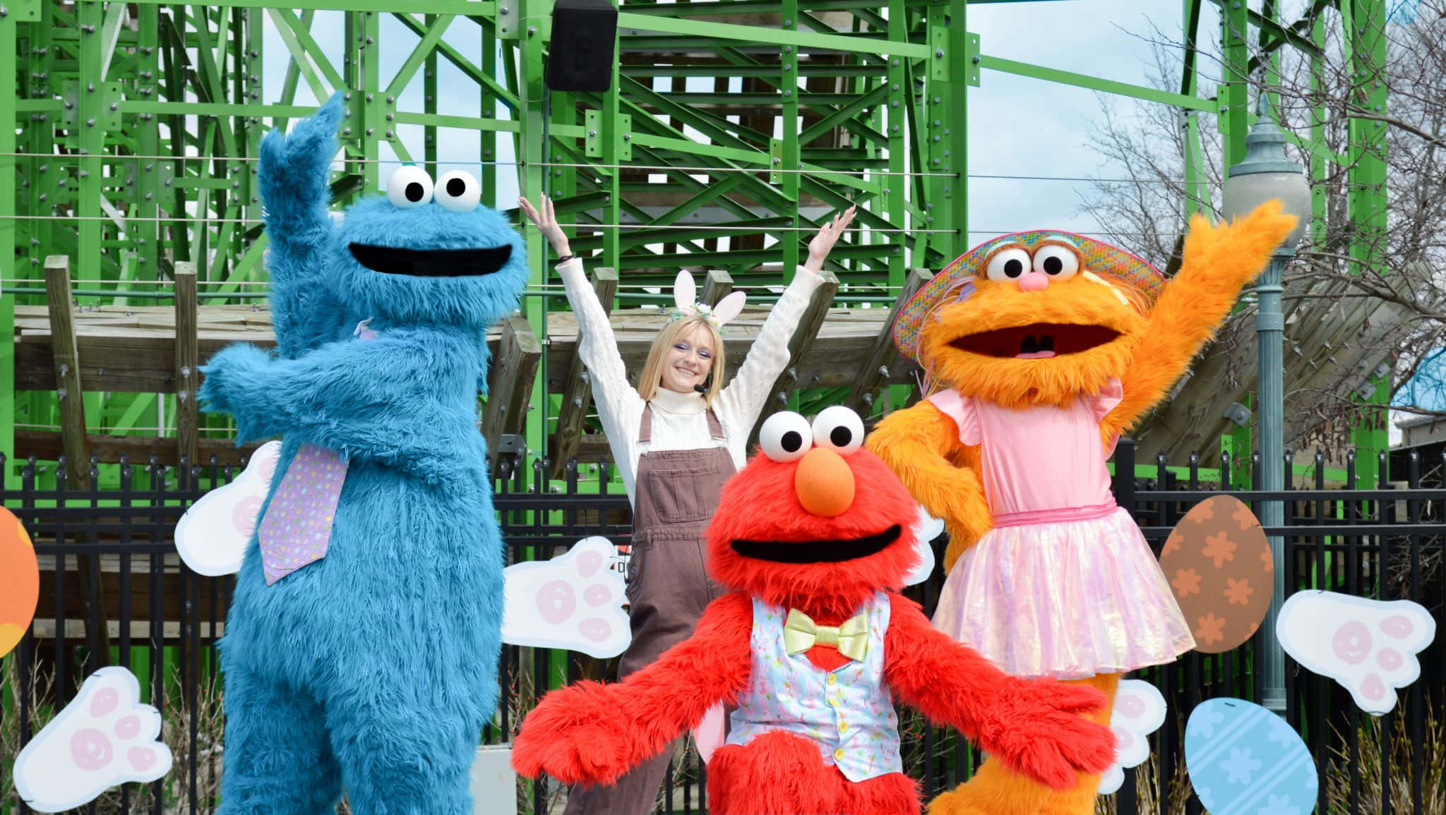 Elmo 桌布 壁紙 — 一位戴著兔耳朵的開朗女士與餅乾怪獸、艾蒙和艾比卡達比站在復活節主題遊樂設施旁。