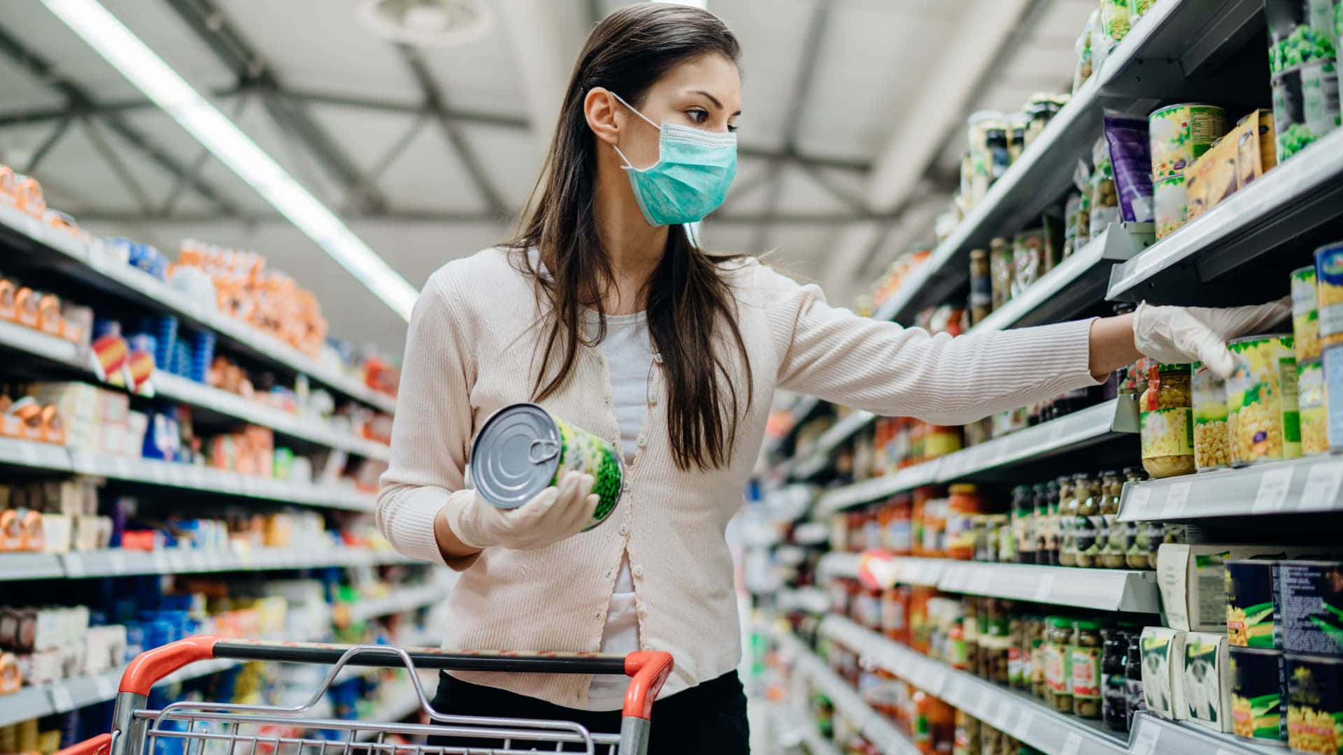 Shopping 桌布 壁紙 — 一位戴著口罩和手套的女性在光線充足的超市通道中挑選罐頭食品。
