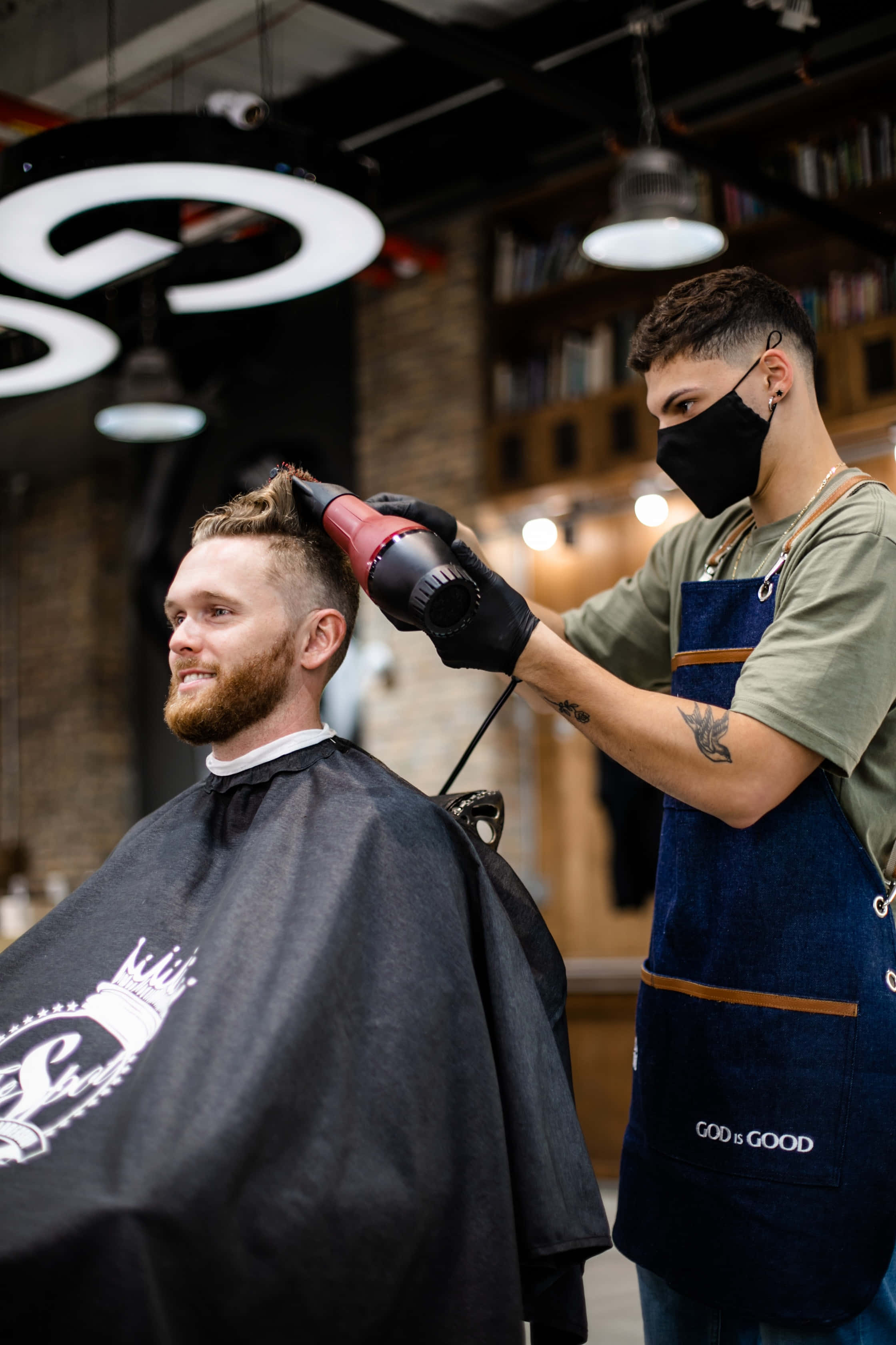 Barber 桌布 壁紙 — 一位戴著口罩和手套的理髮師，在一個有書架和圓形燈具的現代理髮店裡，用紅色吹風機為一位留鬍子的男士造型頭髮。