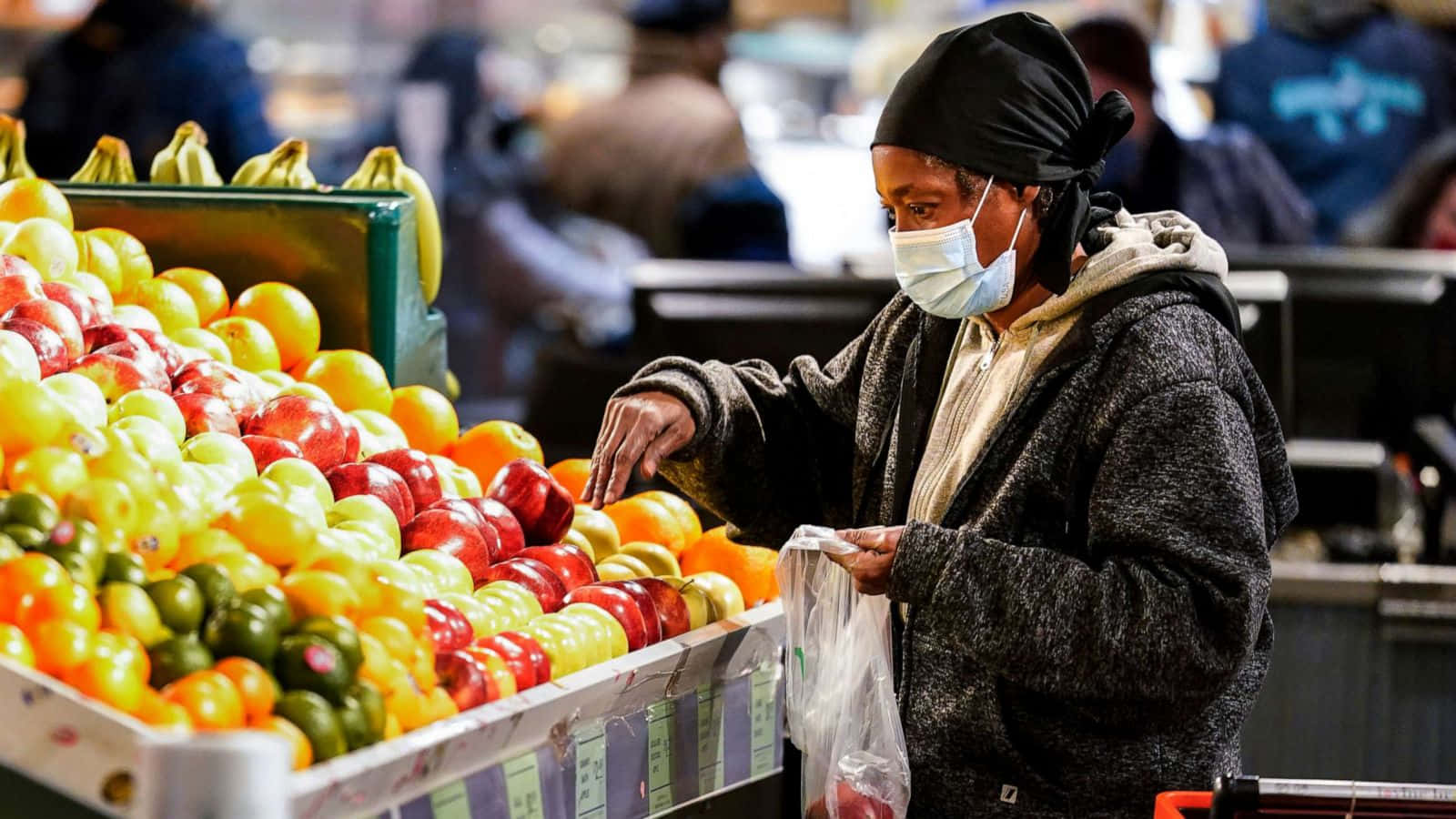 Shopping 桌布 壁紙 — 一位戴著口罩和頭巾的女性在繁忙的室內市場攤位挑選色彩繽紛的番茄。