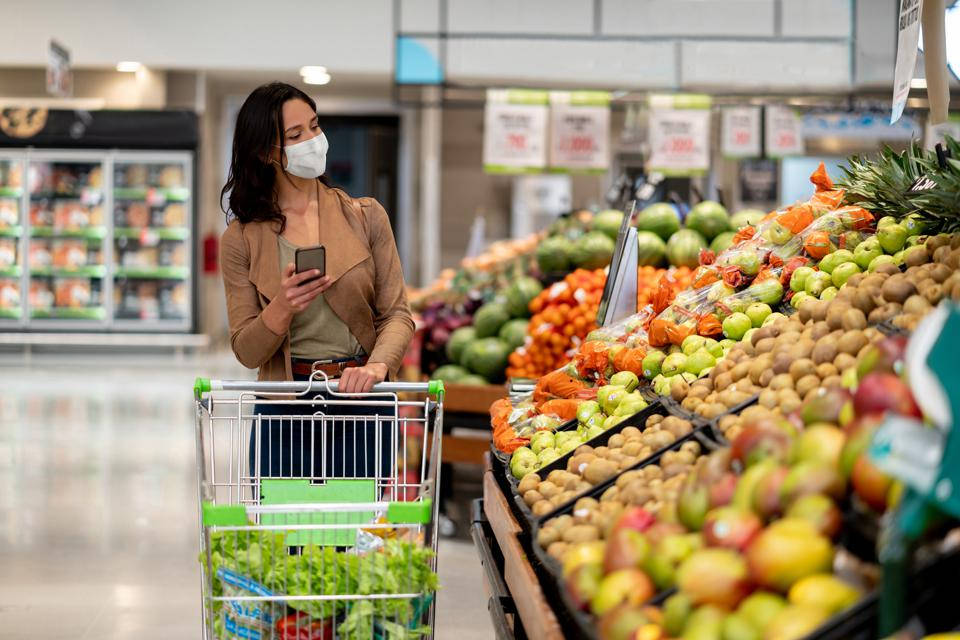 Shopping 桌布 壁紙 — 一位戴著口罩的女性推著購物車，經過光線充足的雜貨店走道中色彩繽紛的農產品陳列。