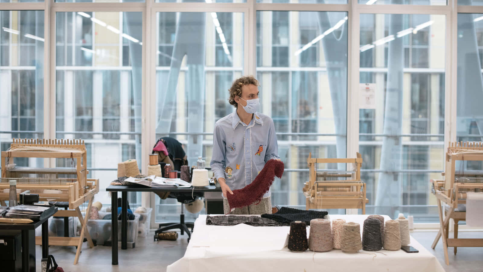 Clothes 桌布 壁紙 — 一位戴著口罩的工匠在明亮現代、充滿織布機和紗線的紡織工作室裡拿著一條紅色針織圍巾。